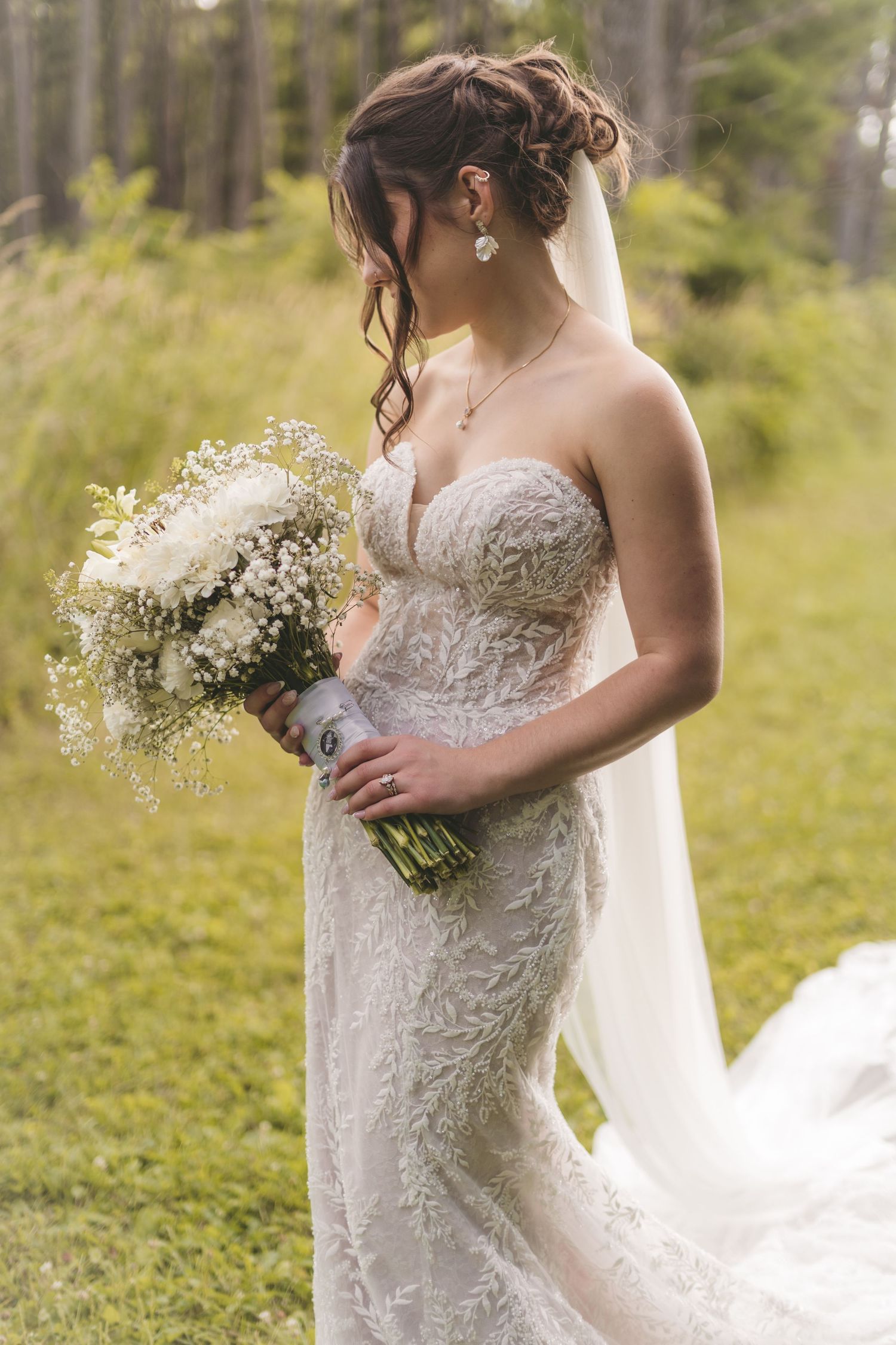 Bride in strapless lace wedding gown holding white floral bouquet outdoors in green forest setting.