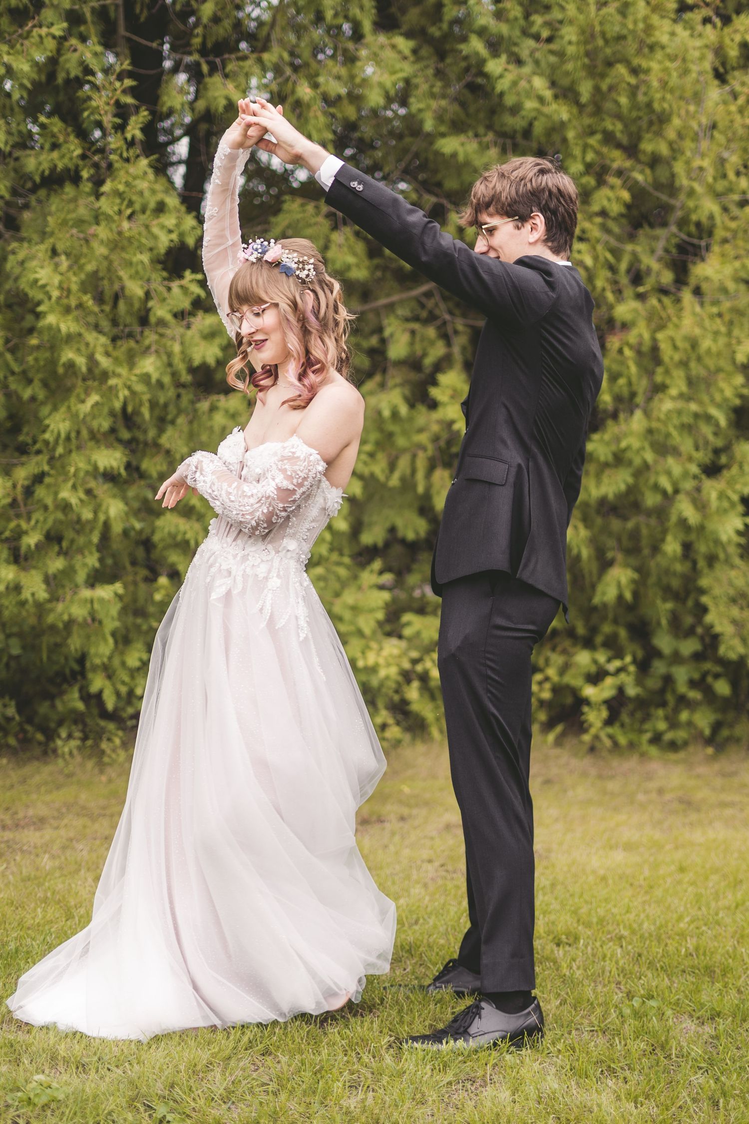 Bride and groom dancing outdoors on grass, groom spinning bride in white lace wedding dress amid green trees.