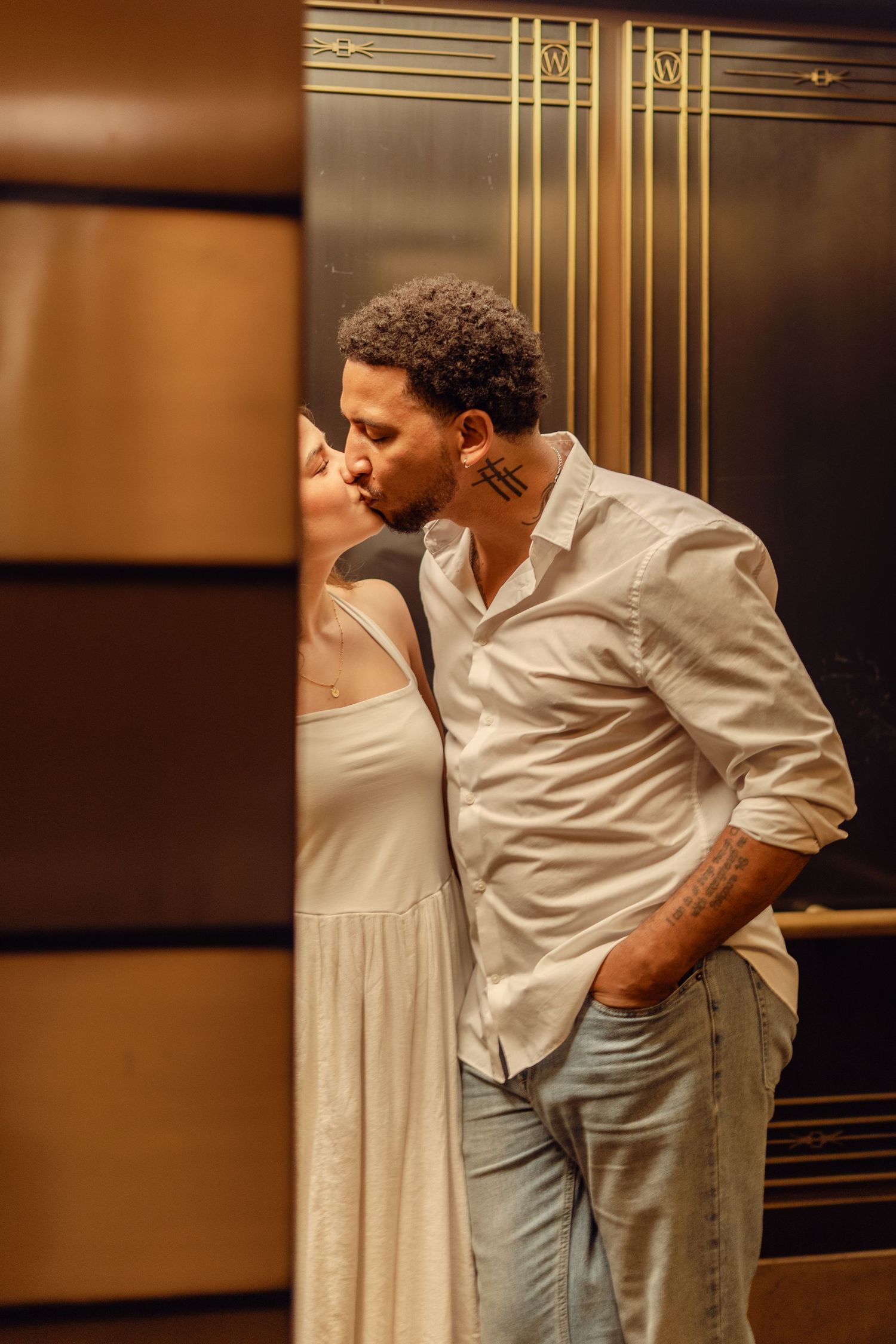 Couple sharing a kiss in front of gold art deco elevator doors, man in white shirt with hand in pocket.