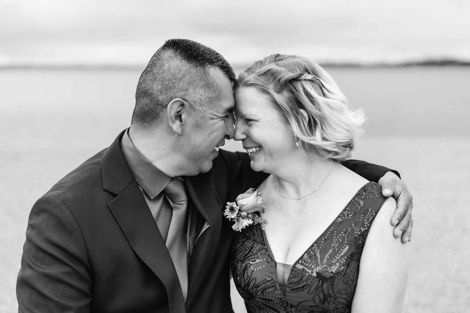 Black and white photo of a smiling couple in formal wear sharing a joyful moment outdoors near water.