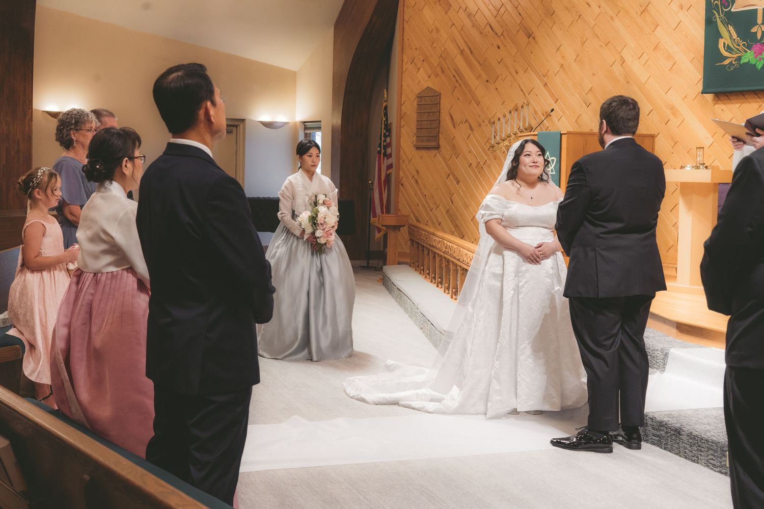 Bride in white gown stands at church altar during wedding ceremony with bridesmaids and guests in background.