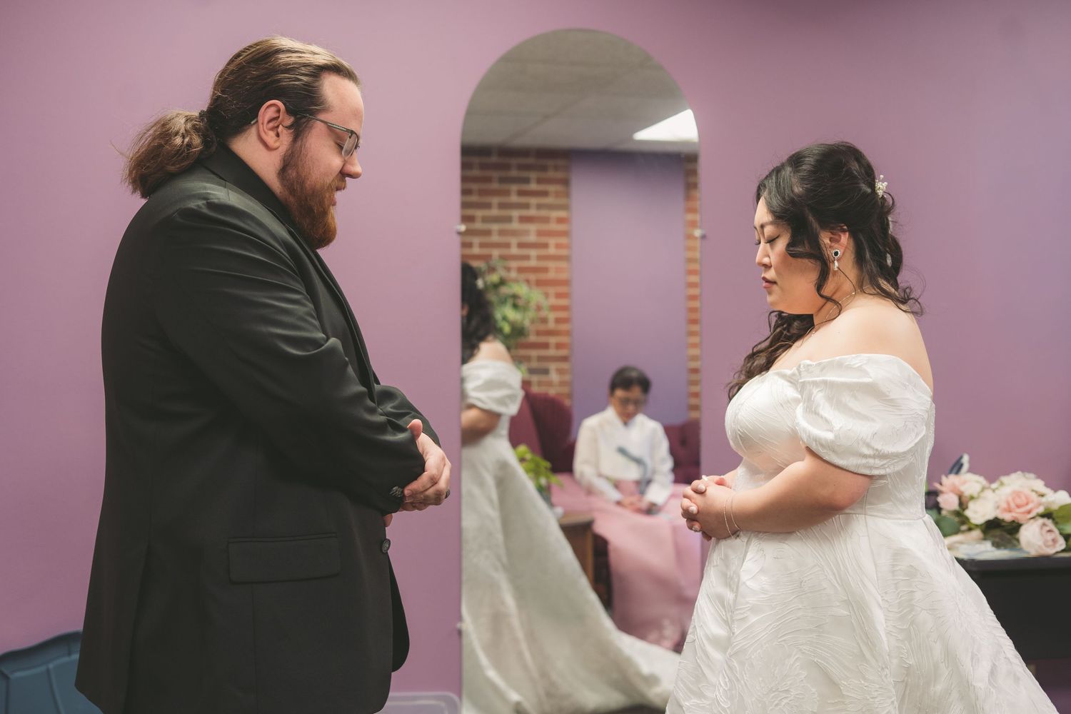 Bride in white off-shoulder gown and groom in black suit share a moment in a purple wedding venue.