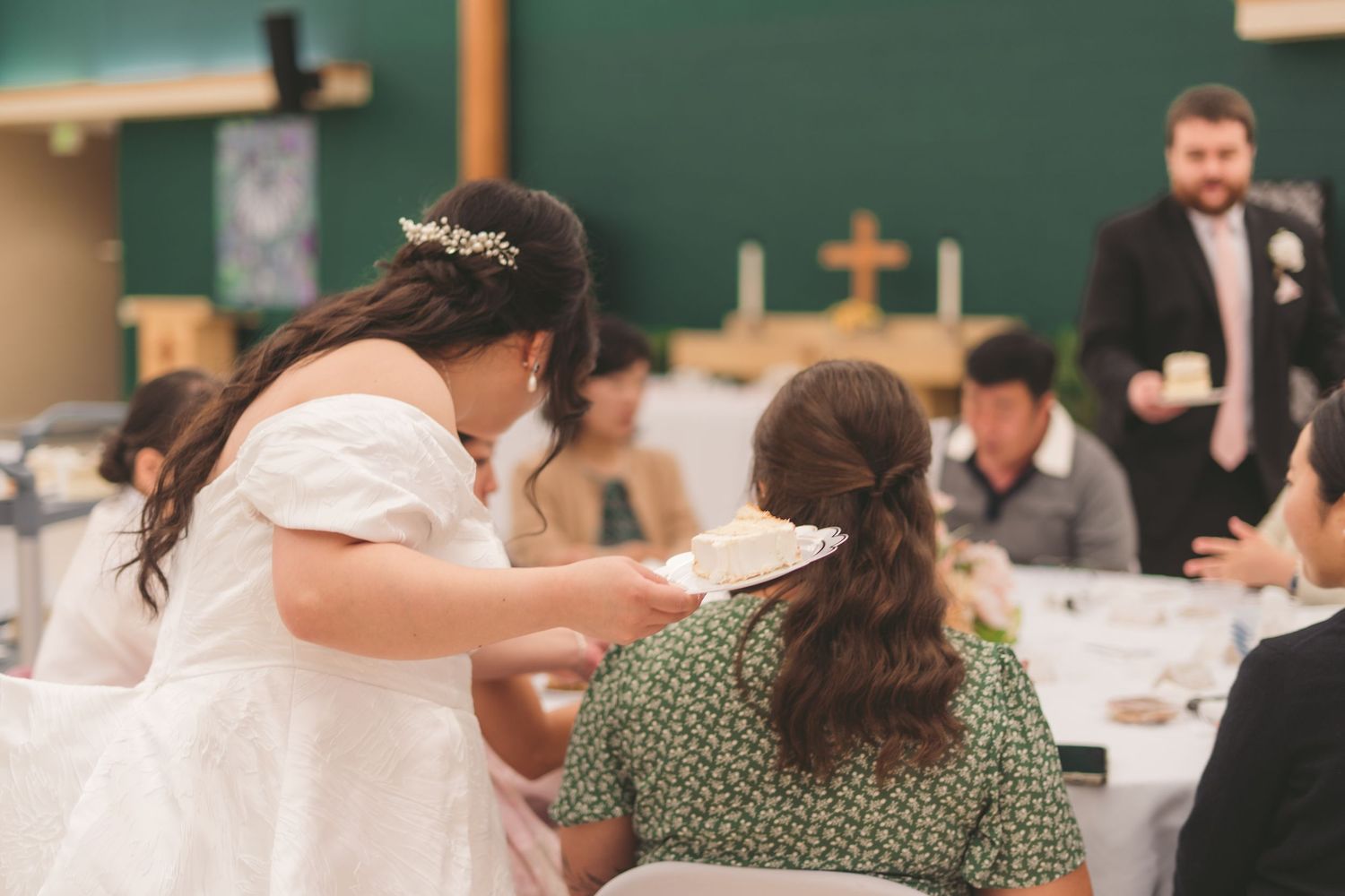 Bride in white gown serves cake to seated guest at elegant wedding reception in green-decorated hall.
