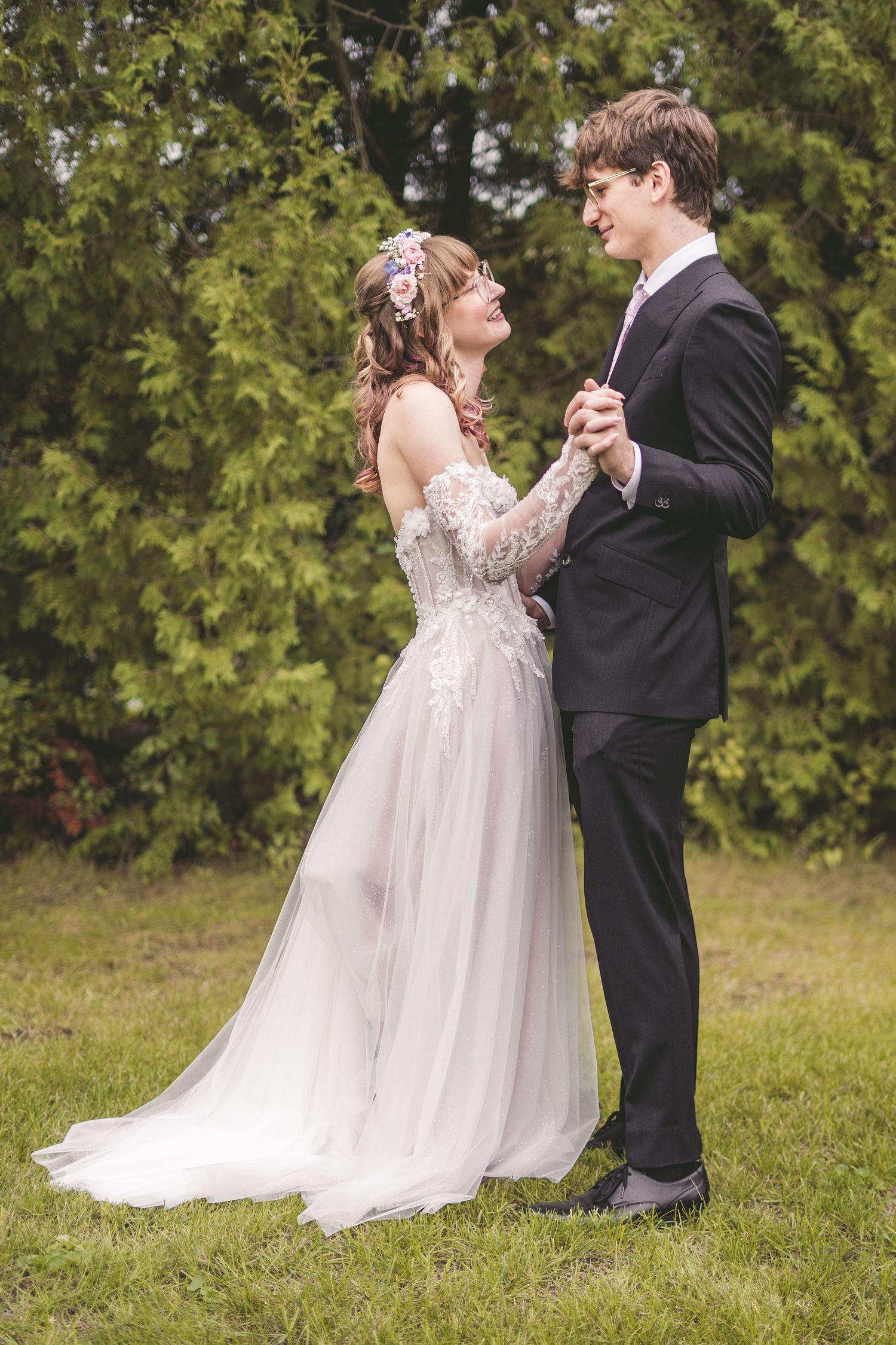 Bride and groom dancing outdoors, she wears a lace off-shoulder gown with floral hair accessories.