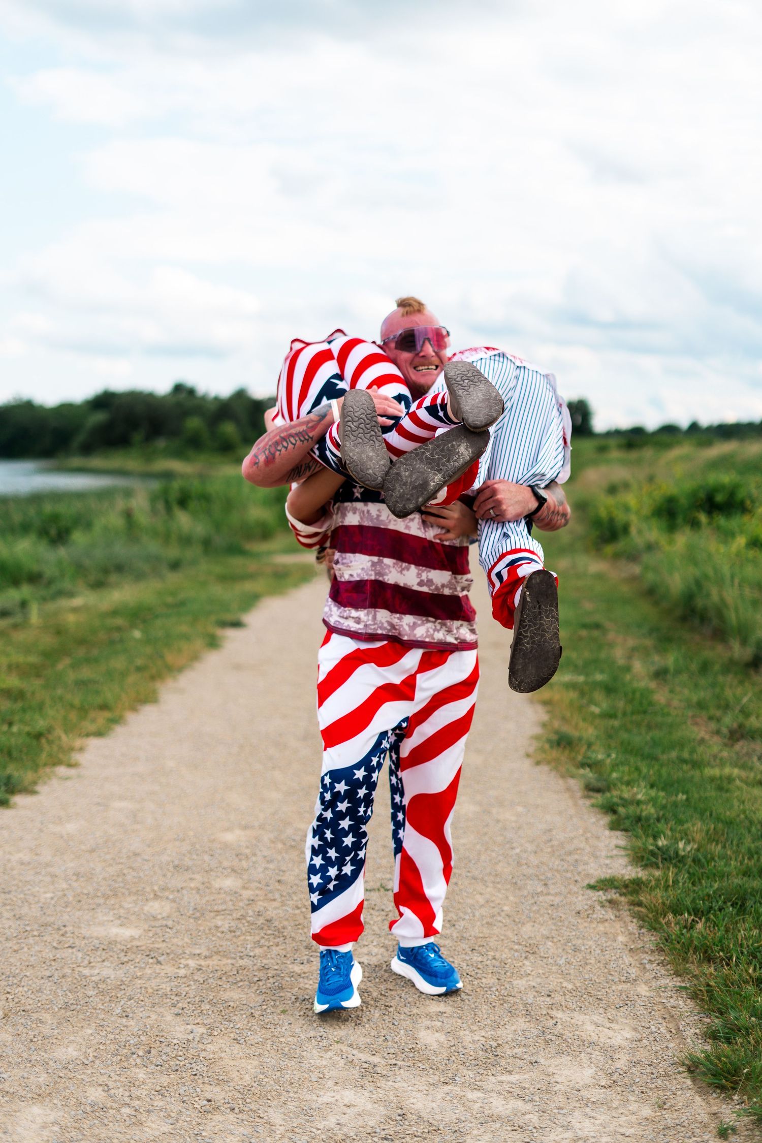 Man in American flag outfit carries another person on a dirt path outdoors.