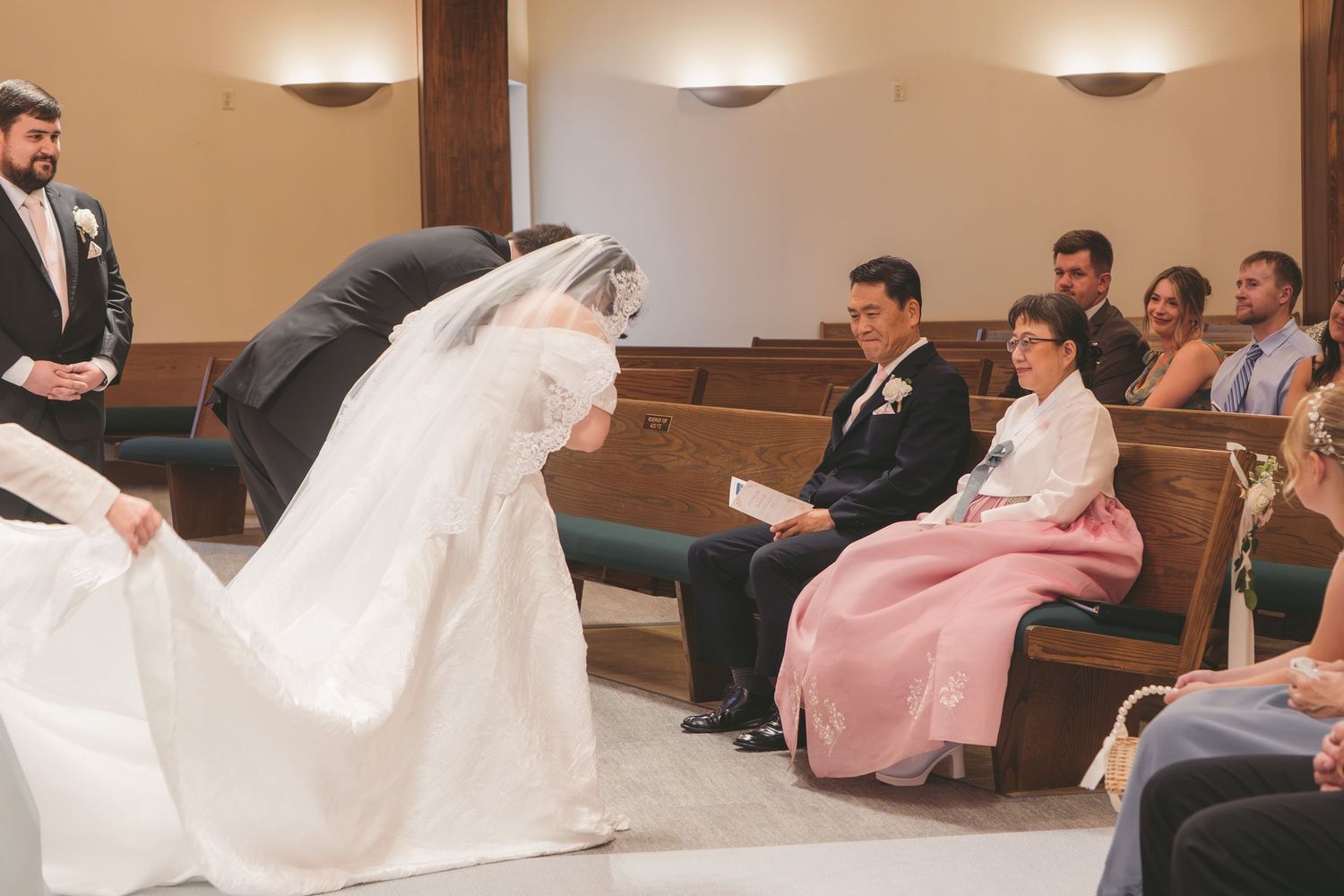 Bride in white gown and veil bows during wedding ceremony in church while guests watch from wooden pews.