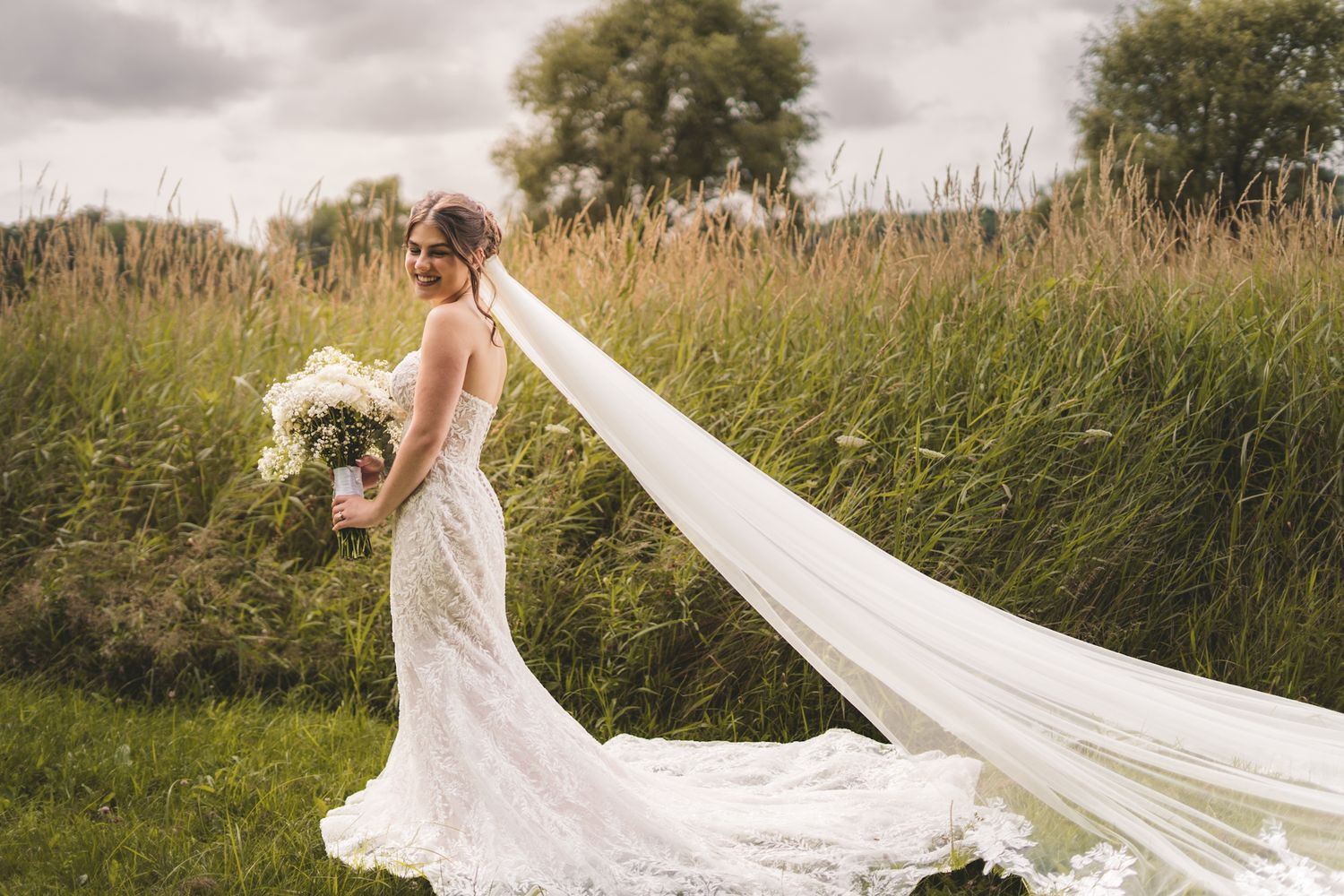 Bride in lace wedding gown with long flowing veil holding bouquet standing in green meadow on cloudy day.