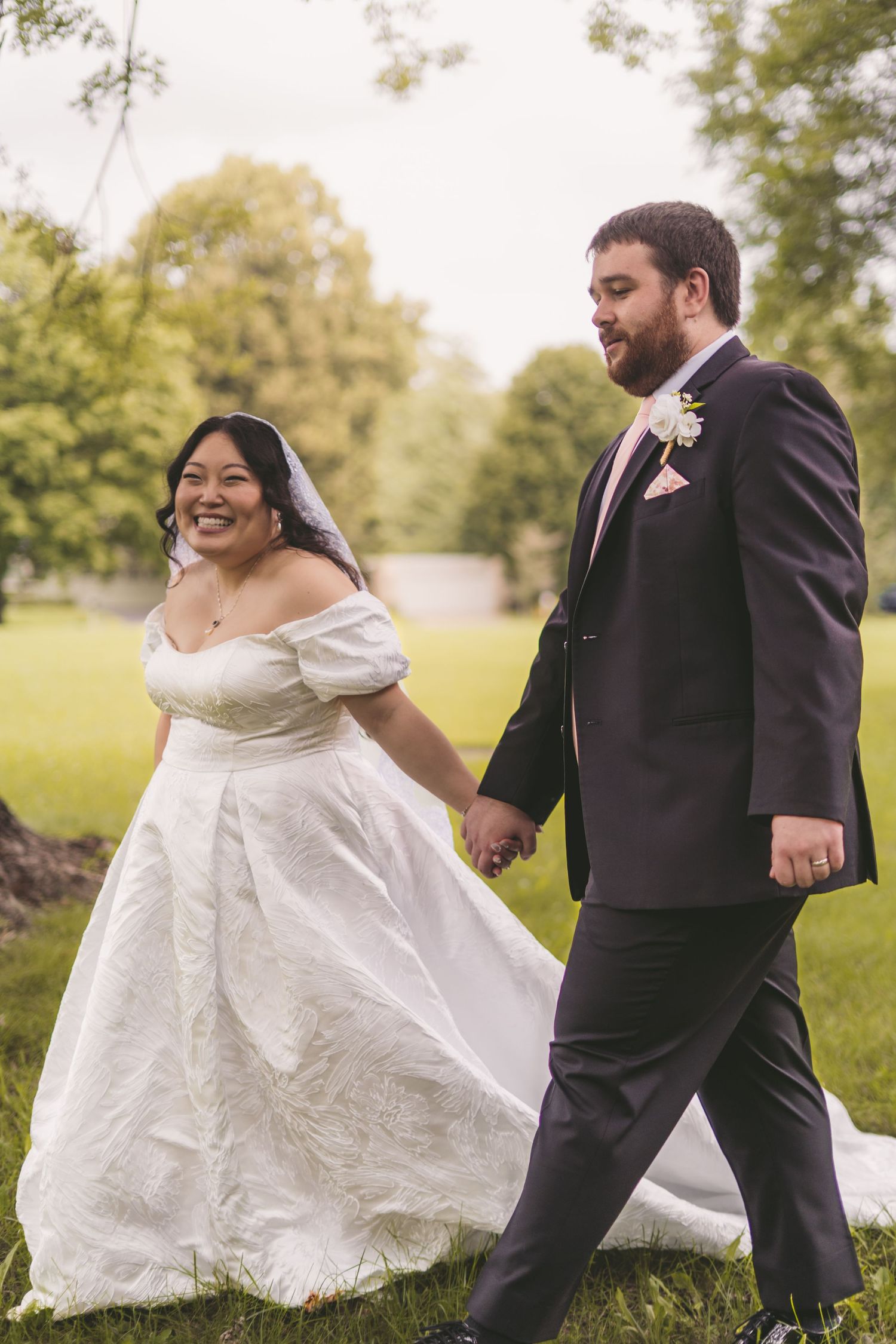 A smiling bride in a white off-shoulder gown and groom in a dark suit hold hands while walking on green grass.