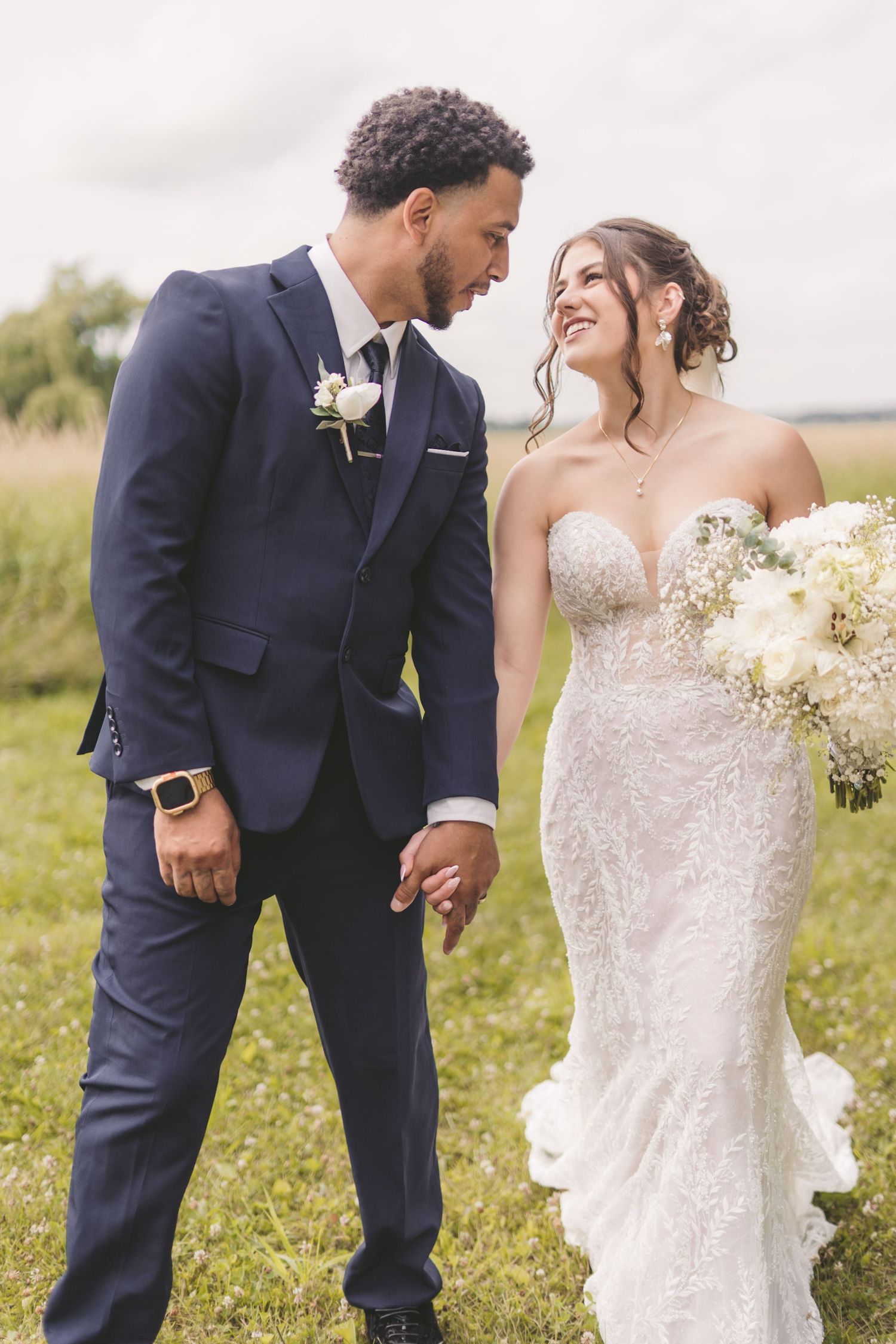 A bride and groom hold hands smiling at each other outdoors, she in a lace gown, he in a navy suit.