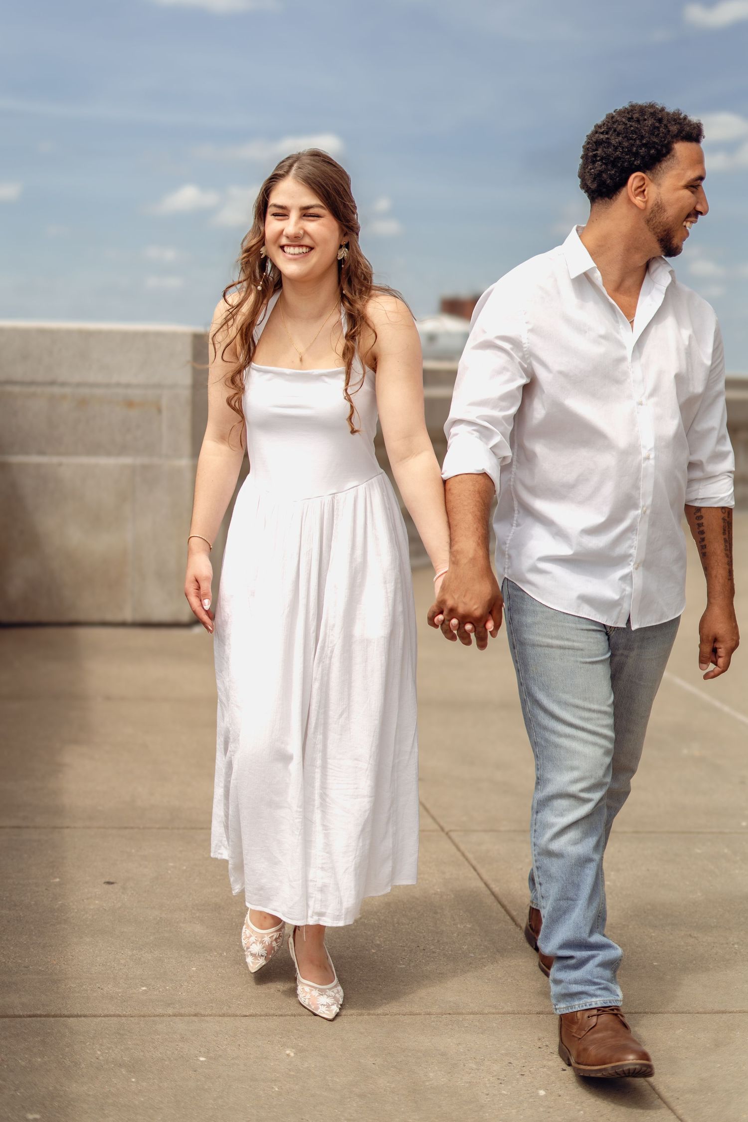 Smiling couple holding hands walking on rooftop, woman in white maxi dress and man in white shirt with blue jeans.