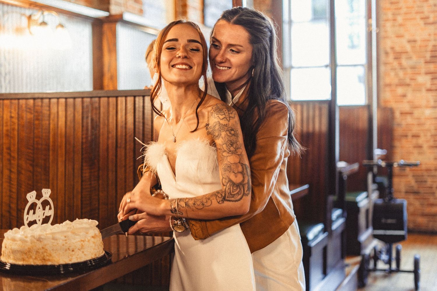 Two brides share a joyful moment together while cutting their wedding cake in a rustic venue with brick walls.