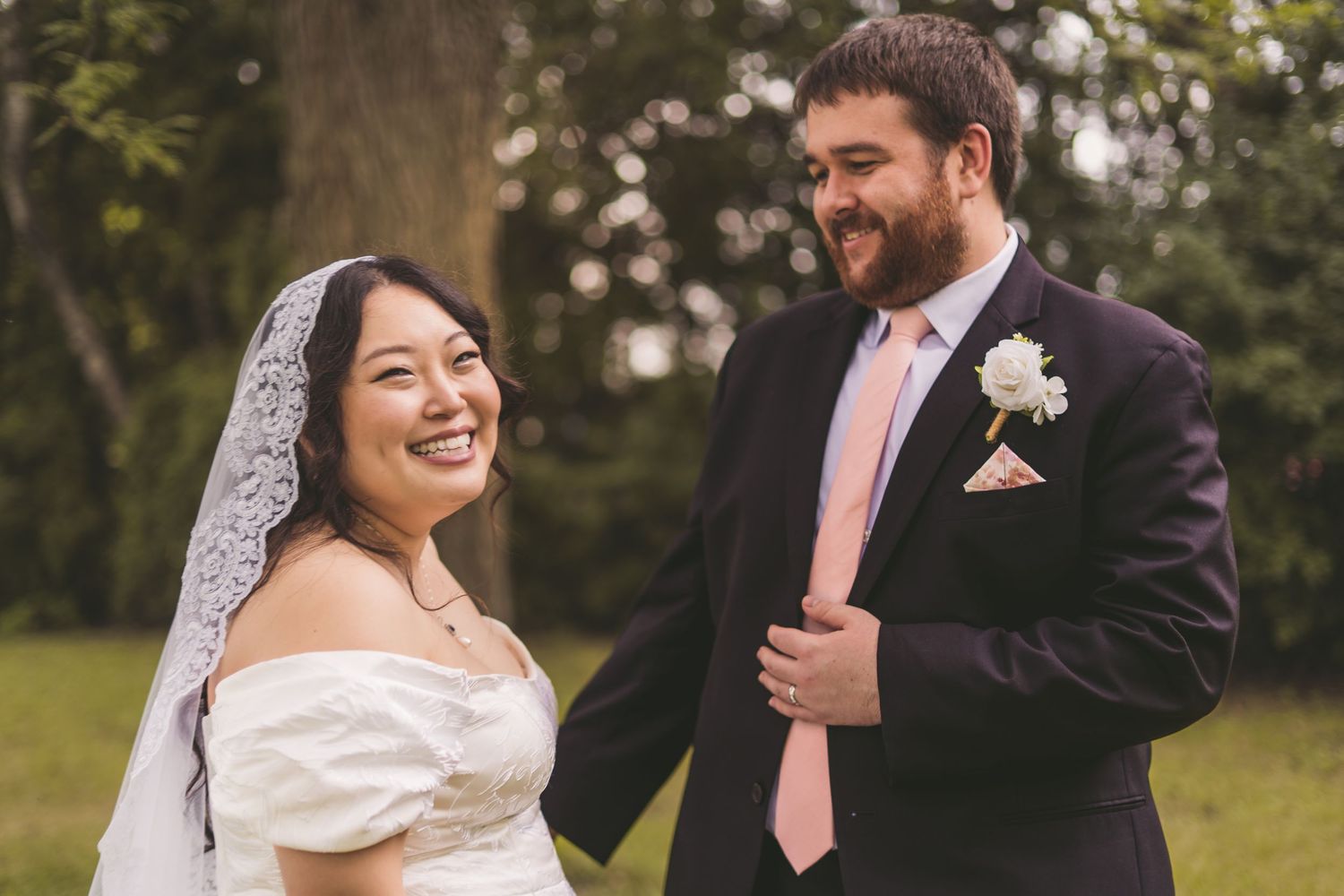 A smiling bride in a white off-shoulder gown and veil stands with a groom in a navy suit and pink tie outdoors.