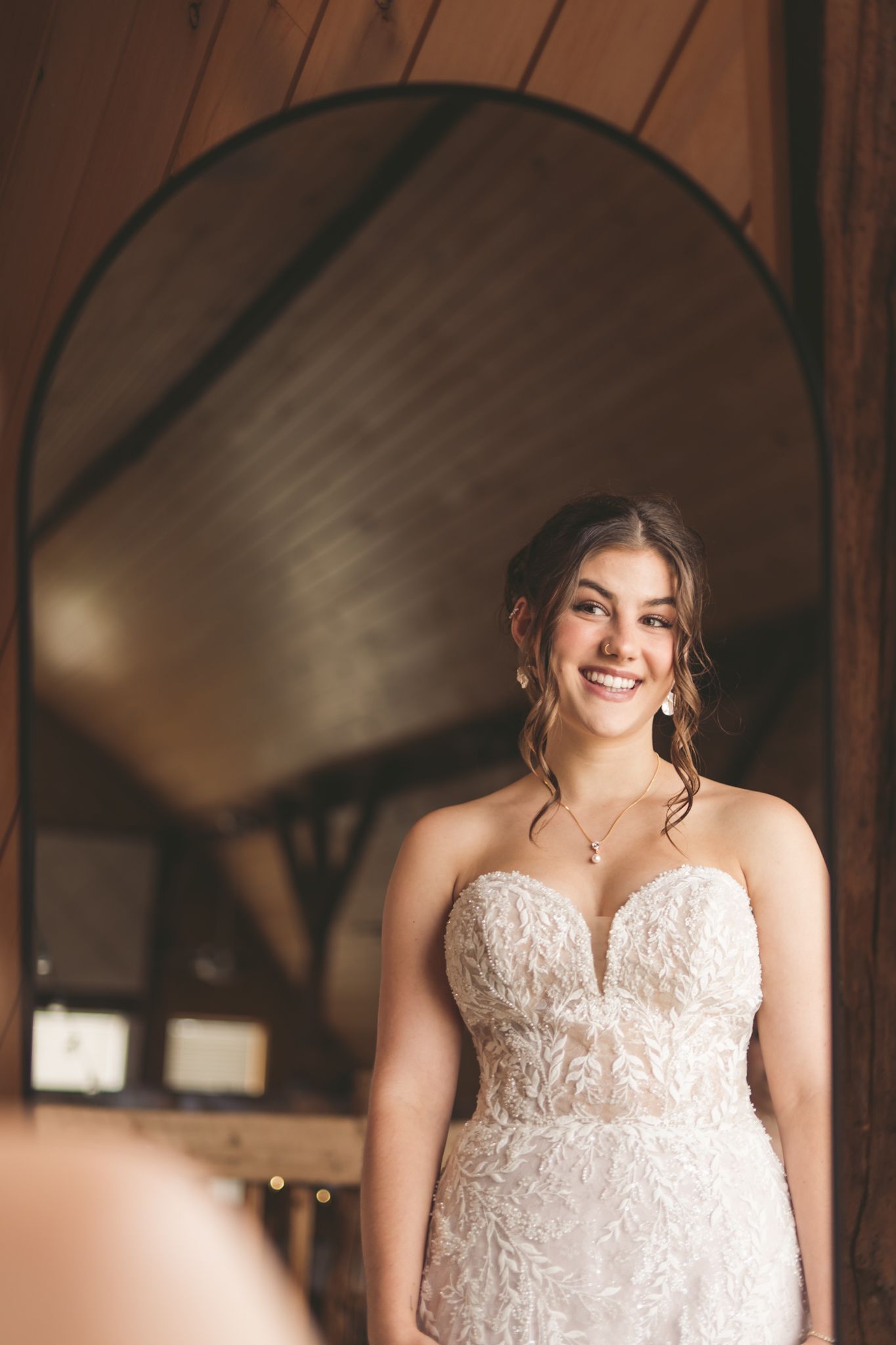 Smiling bride in a strapless lace wedding gown standing before an arched mirror in a rustic barn venue.