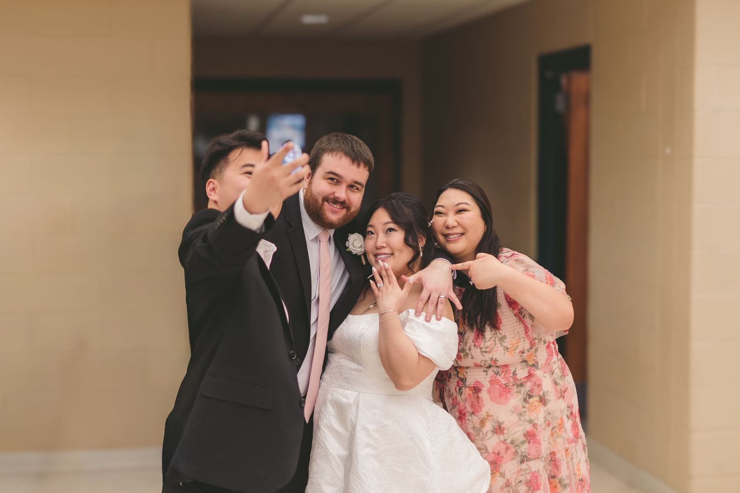 Wedding group selfie with bride in white dress and groom in suit posing with two guests in a hallway.