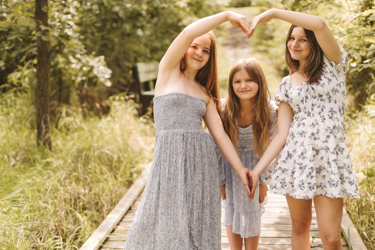 Three girls pose on a wooden path in nature, two forming a heart shape with their arms overhead.