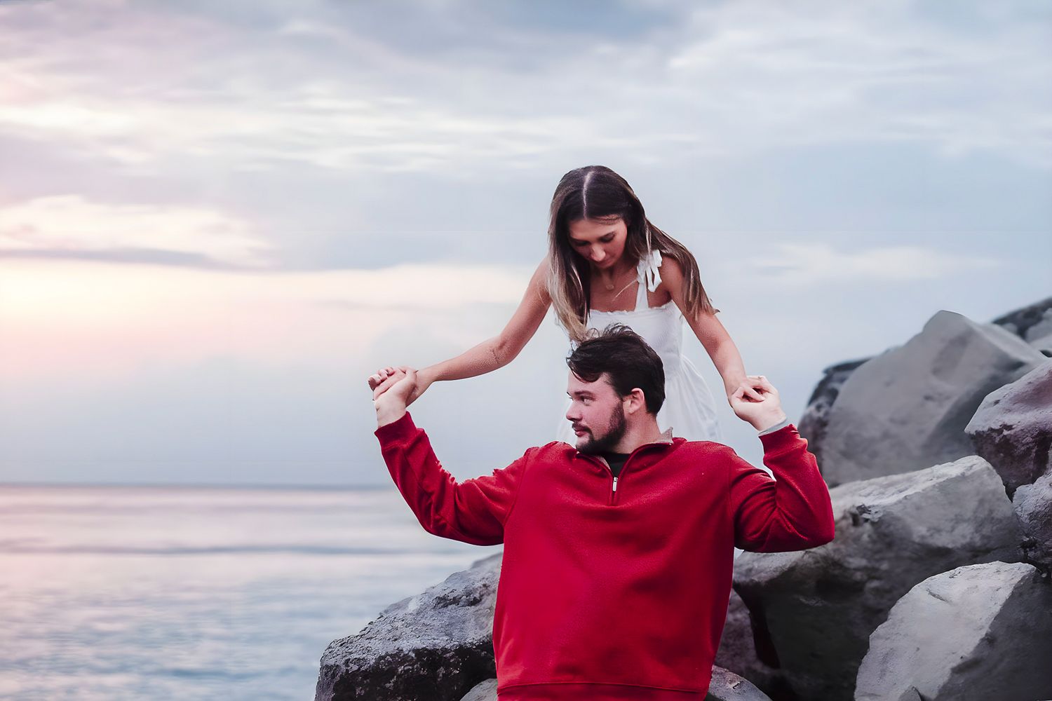 A couple poses romantically on rocky shore at sunset, woman standing behind seated man holding his hands up.