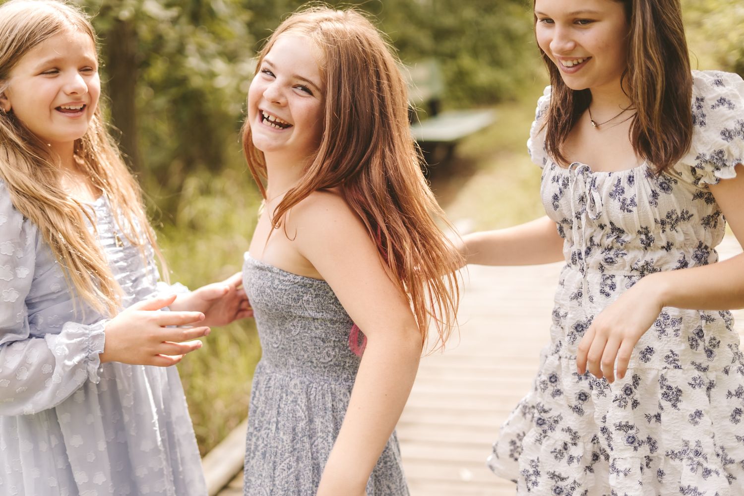 Three girls laughing and playing outdoors in summer dresses on a wooden bridge surrounded by nature.