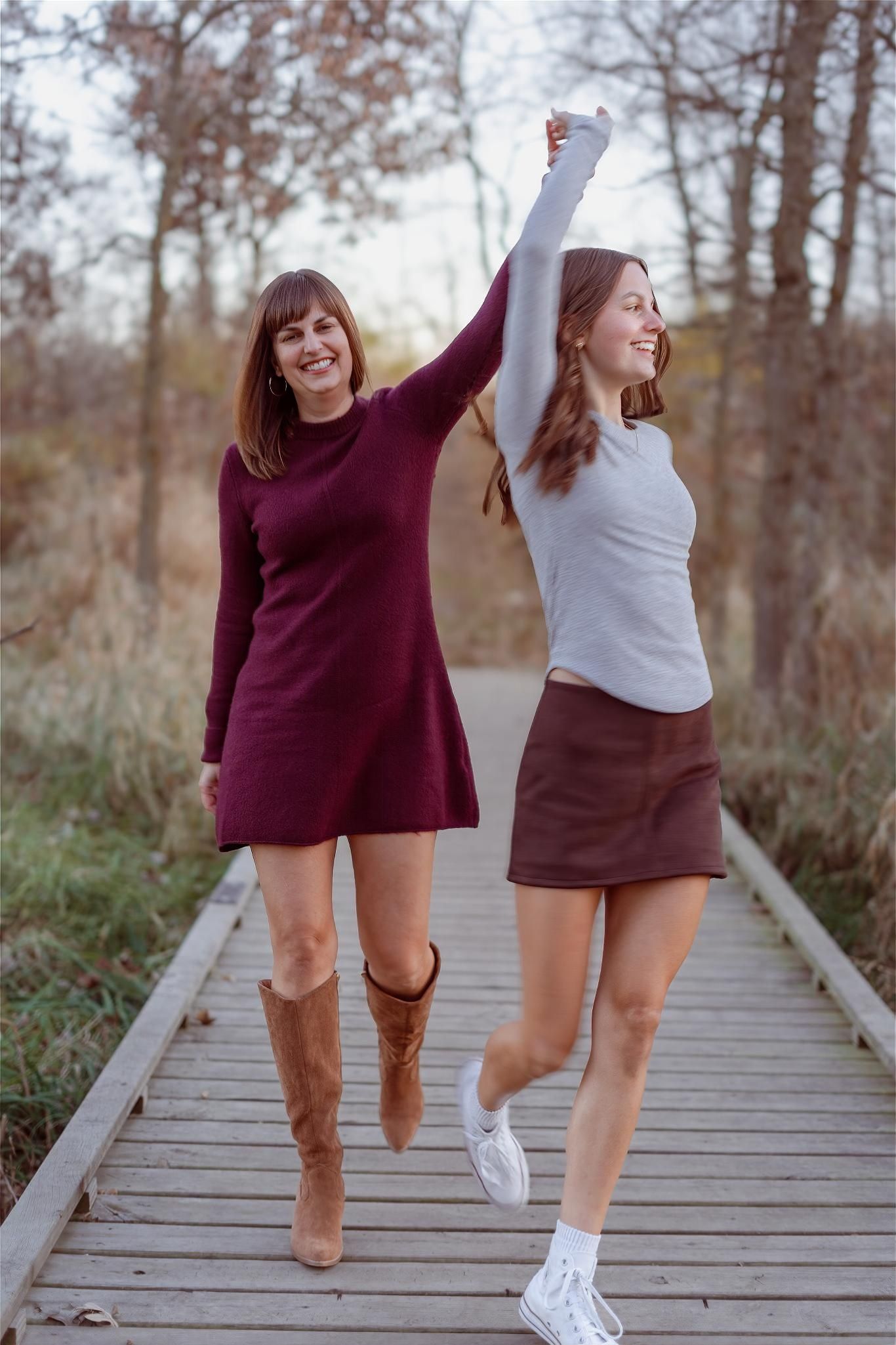 Two happy women holding hands on a wooden boardwalk path in autumn, wearing stylish dresses and boots.