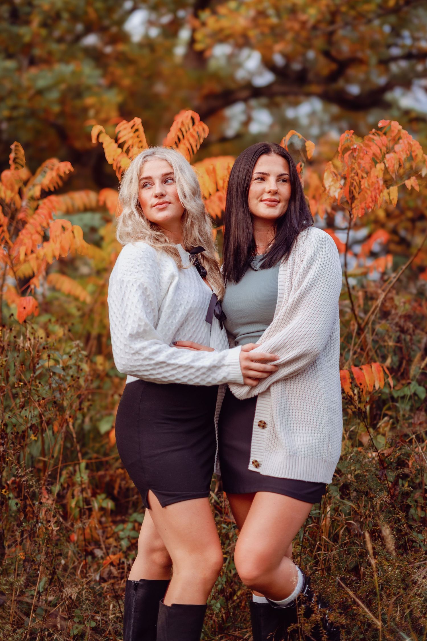 Two sisters in sweaters and boots pose against vibrant fall foliage.