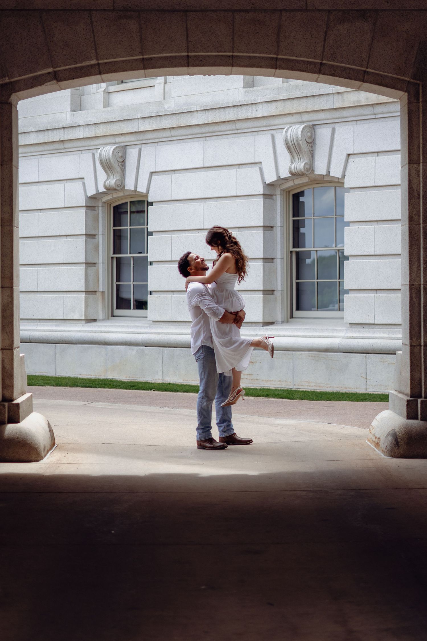 Man lifting woman in white dress under stone archway outside classic white marble building, sharing a kiss.