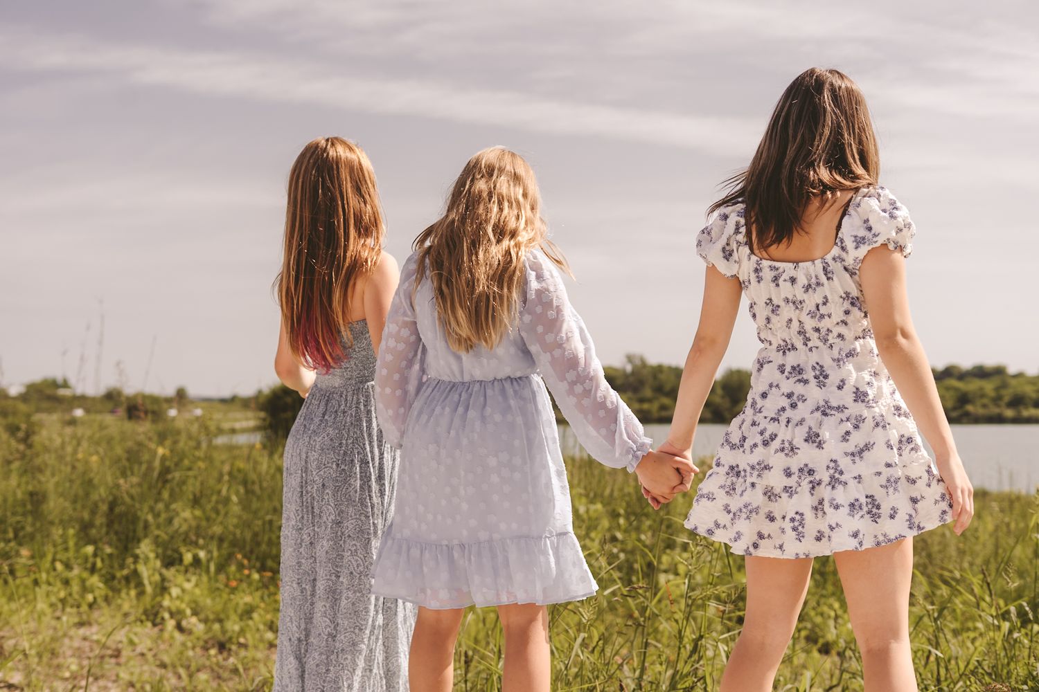 Three young women in floral dresses holding hands while walking through a green field near a lake.