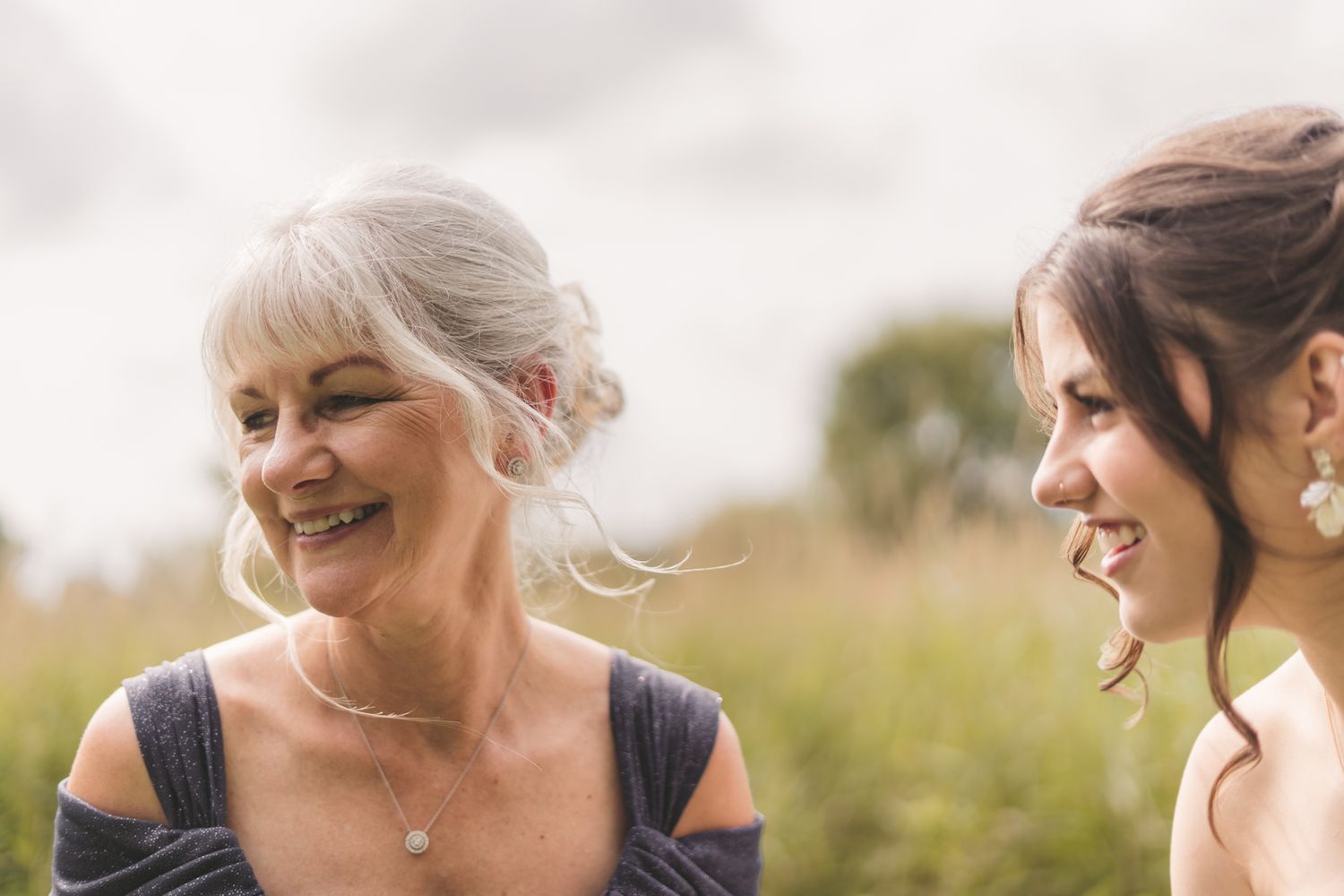 Two women smiling outdoors at a wedding, older woman with silver updo and younger brunette bride in elegant attire.