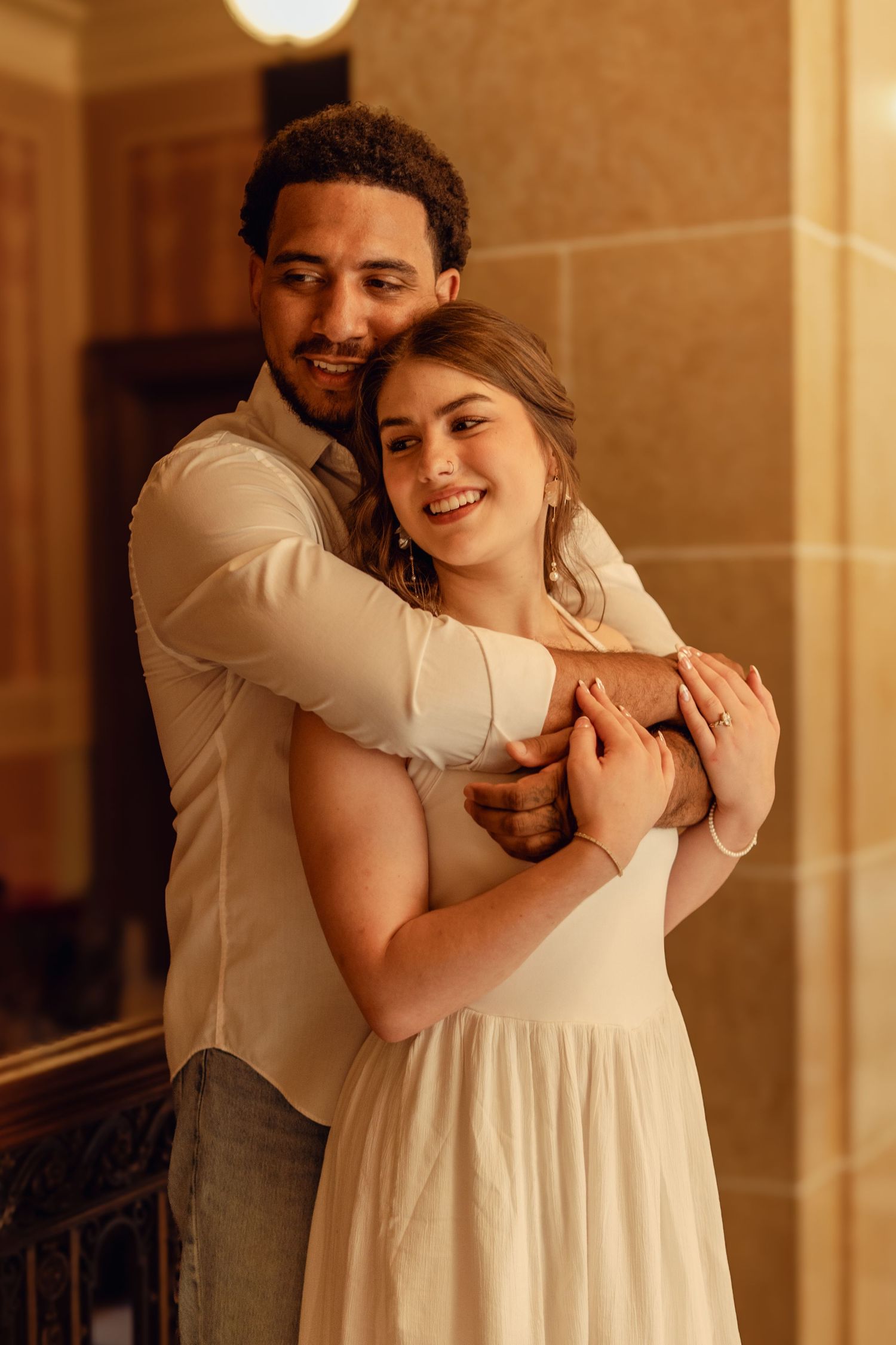 A couple embraces warmly in golden light, the man hugging the smiling woman in a white dress from behind.