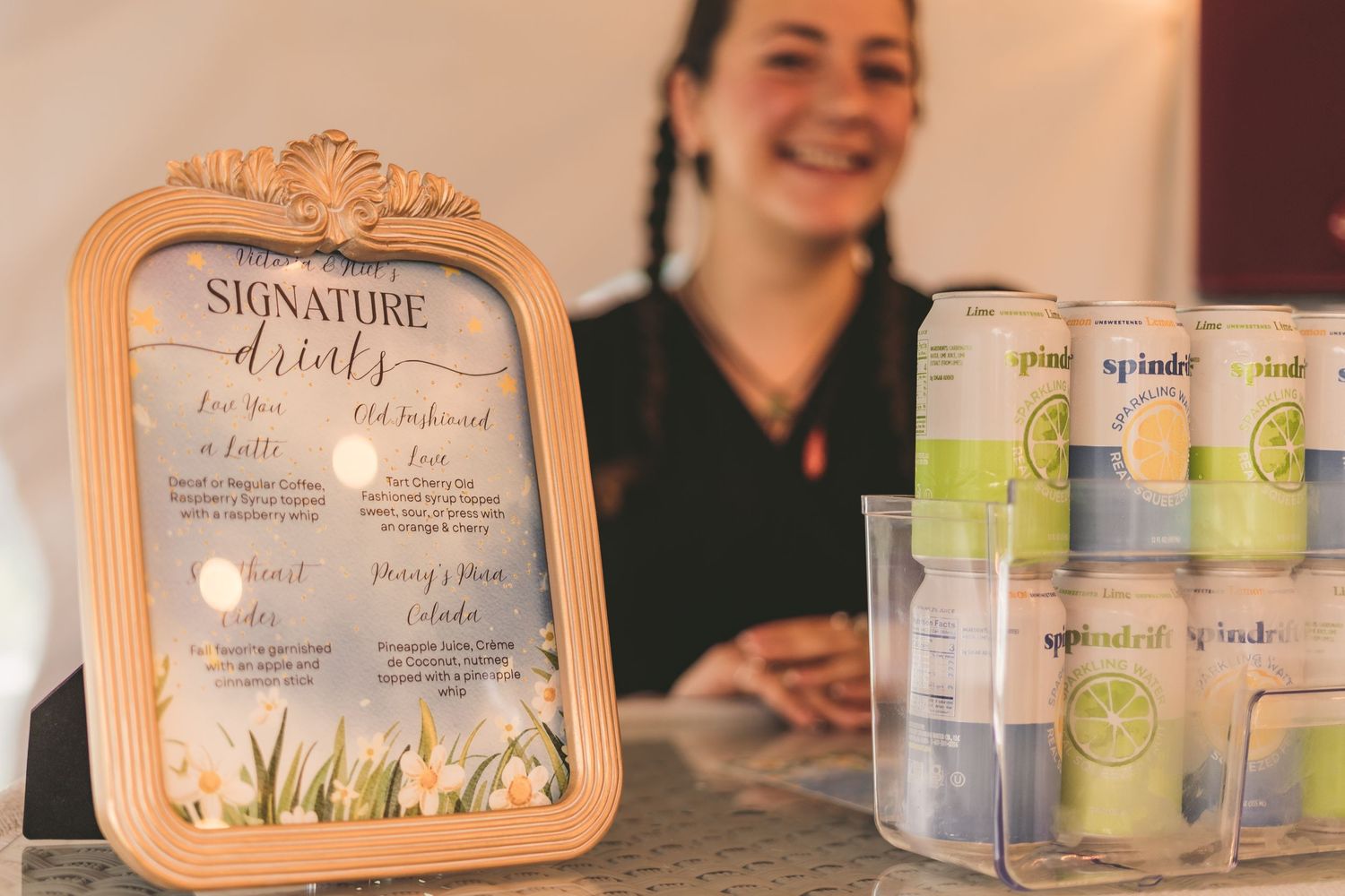 Bartender smiling behind a signature drinks menu sign with Spindrift sparkling water cans at a wedding bar.