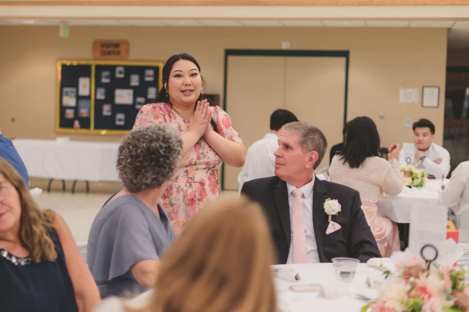 Woman in floral dress clapping at elegant wedding reception dinner with guests seated at decorated tables.