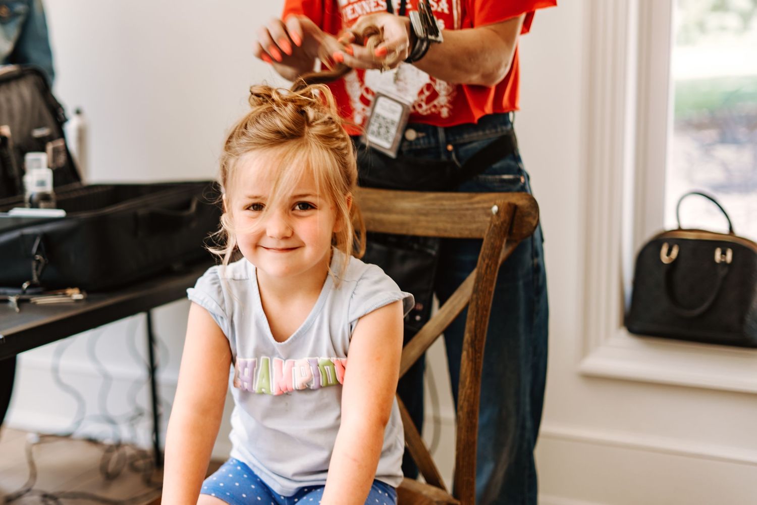 Young girl smiling while getting her hair styled, sitting on a wooden chair indoors.