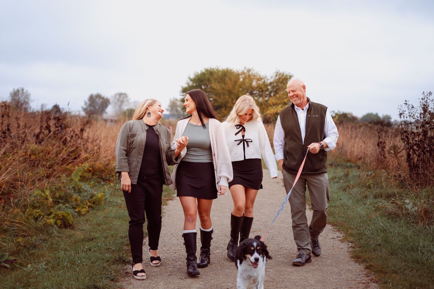 Family of four walking a border collie dog on a scenic autumn trail path surrounded by golden fields.