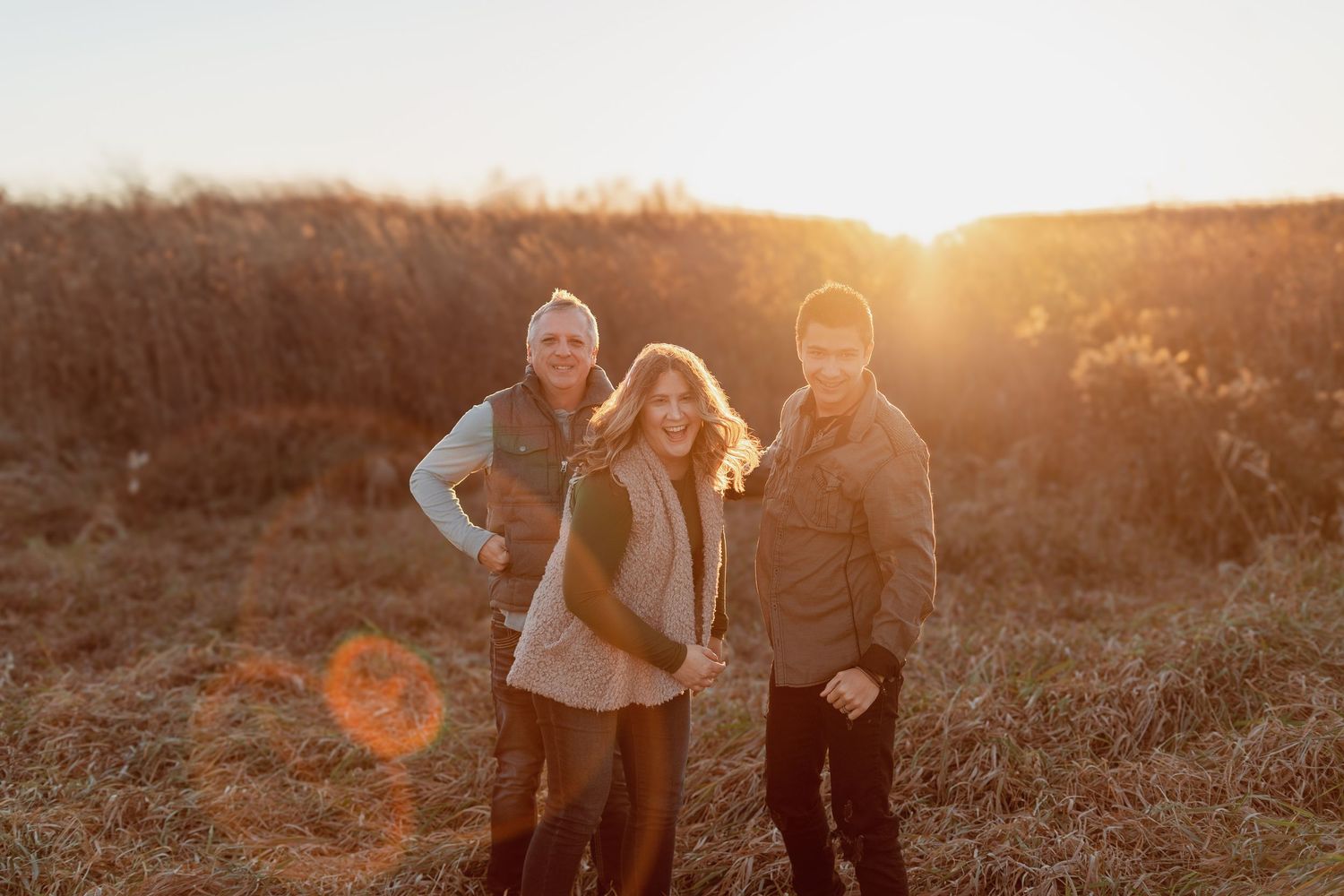 Three people smiling outdoors at golden hour, standing in a field with warm sunlight glowing behind them.