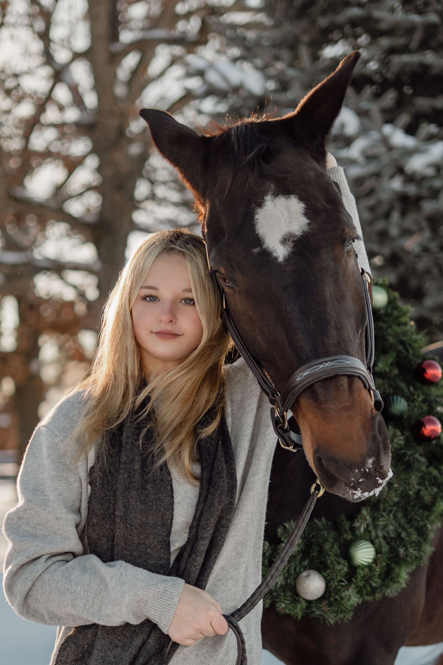 Young blonde woman posing with a dark horse wearing a Christmas wreath in a snowy winter outdoor setting.