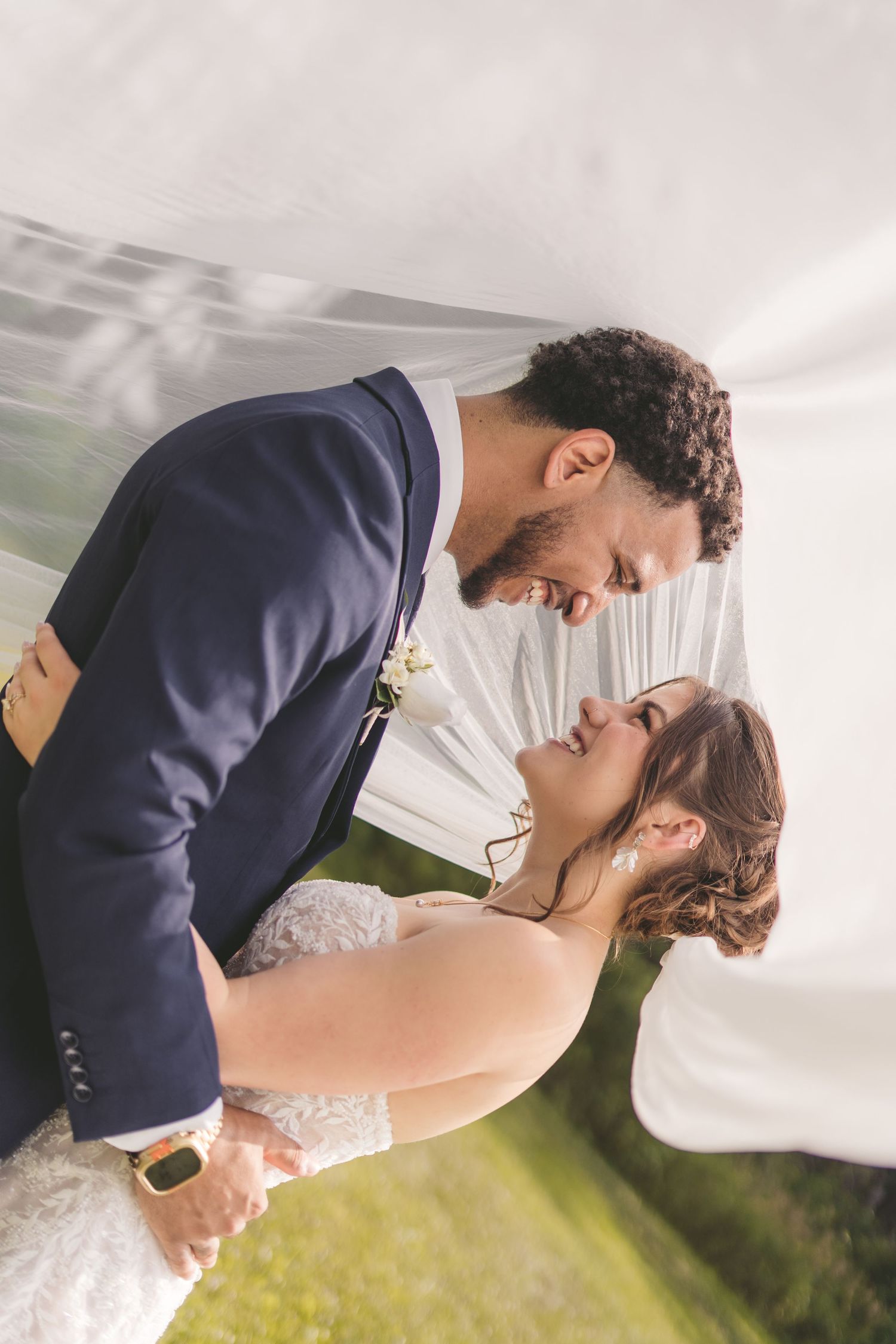 Bride and groom sharing a romantic moment under a flowing white veil outdoors on their wedding day.