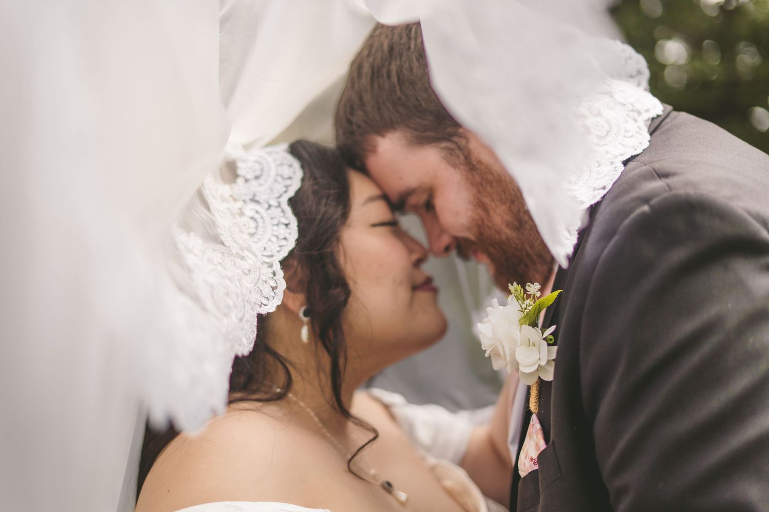 Bride and groom share romantic kiss under lace wedding veil outdoors on their wedding day.