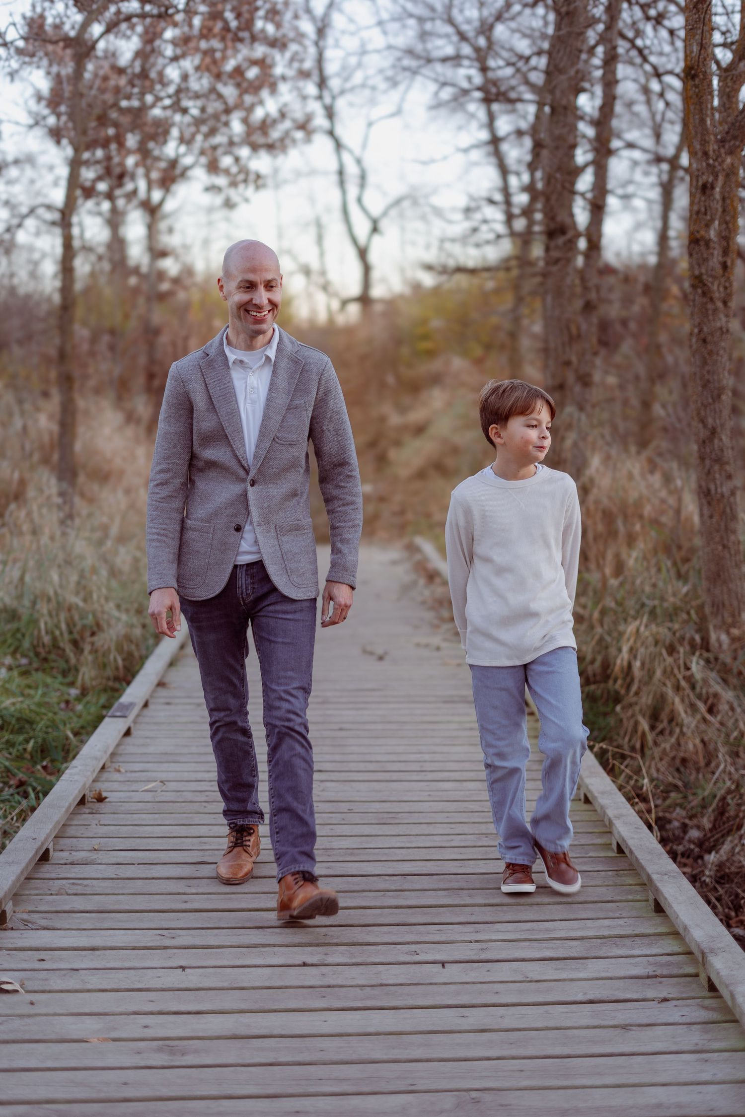 A smiling man in a gray blazer and a young boy walk together on a wooden boardwalk path through autumn trees.