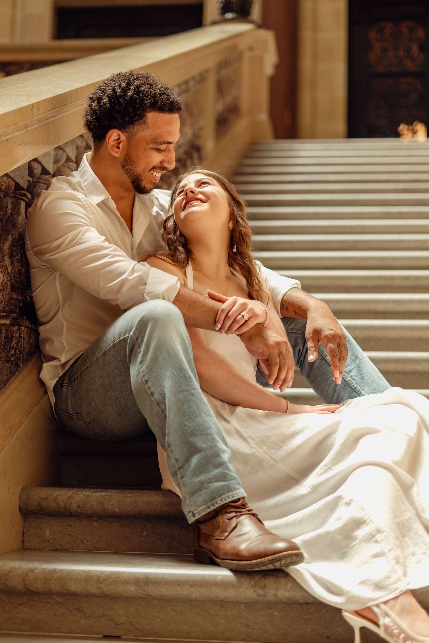 Happy interracial couple sitting on stone steps, laughing and embracing in warm golden light.