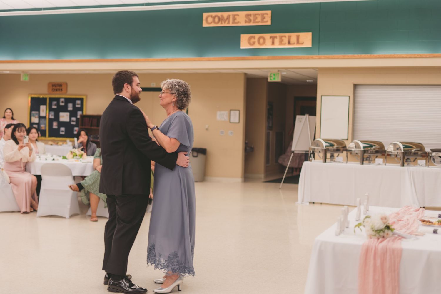 Couple sharing a mother-son dance at a wedding reception in a decorated hall with guests seated nearby.