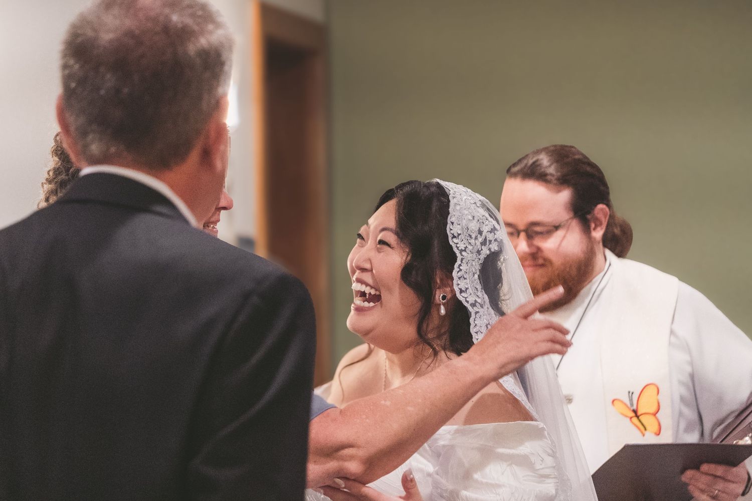 Bride laughing joyfully with groom during wedding ceremony, officiant facing couple at altar.