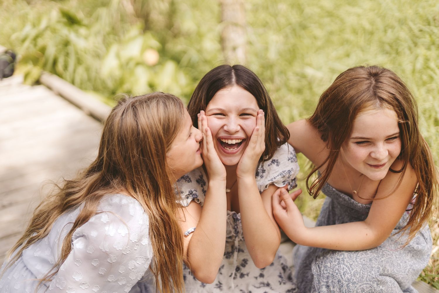 Three girls laughing and whispering secrets outdoors in a sunny green park setting.