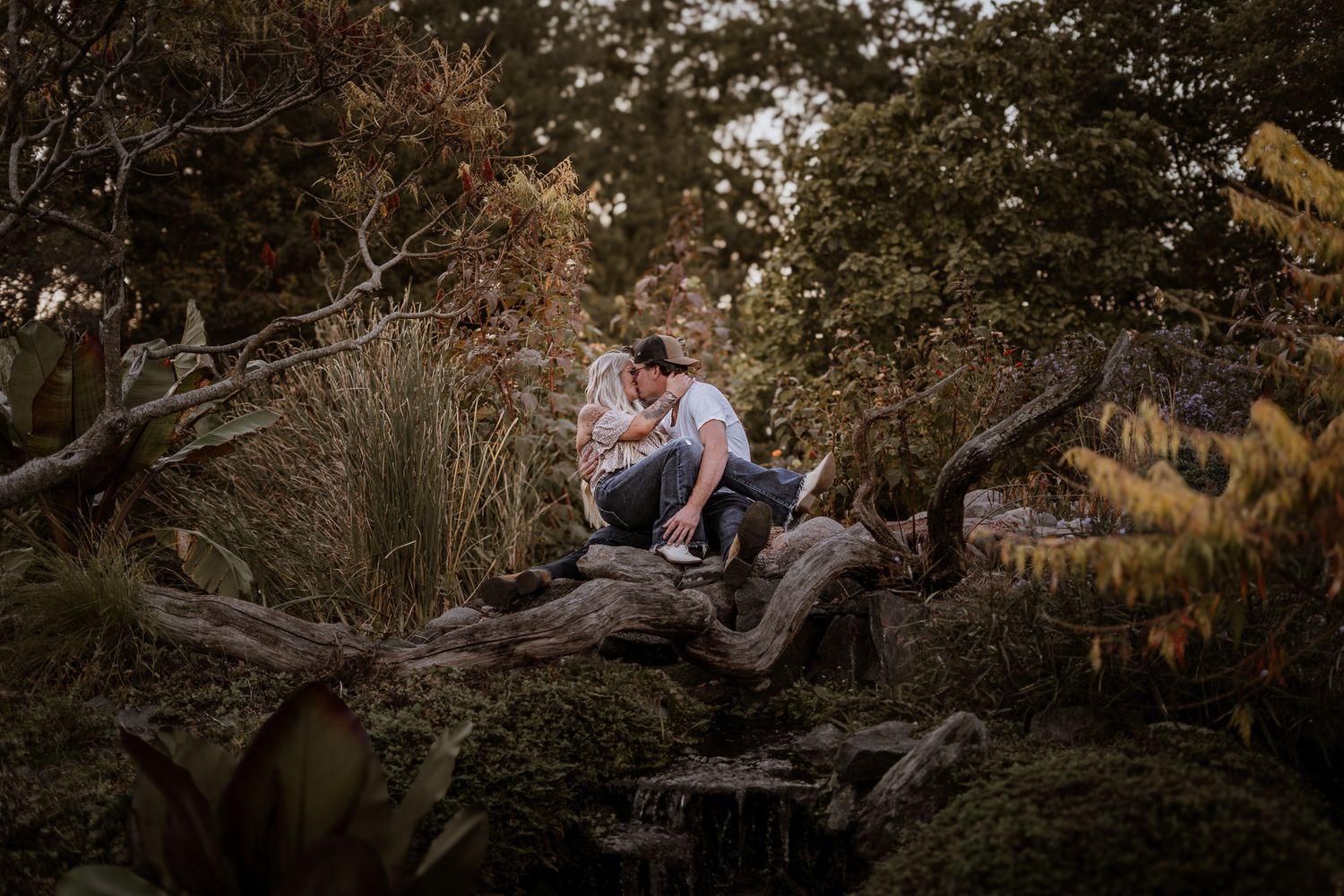 A couple sits together on fallen logs in a wooded area at sunset.