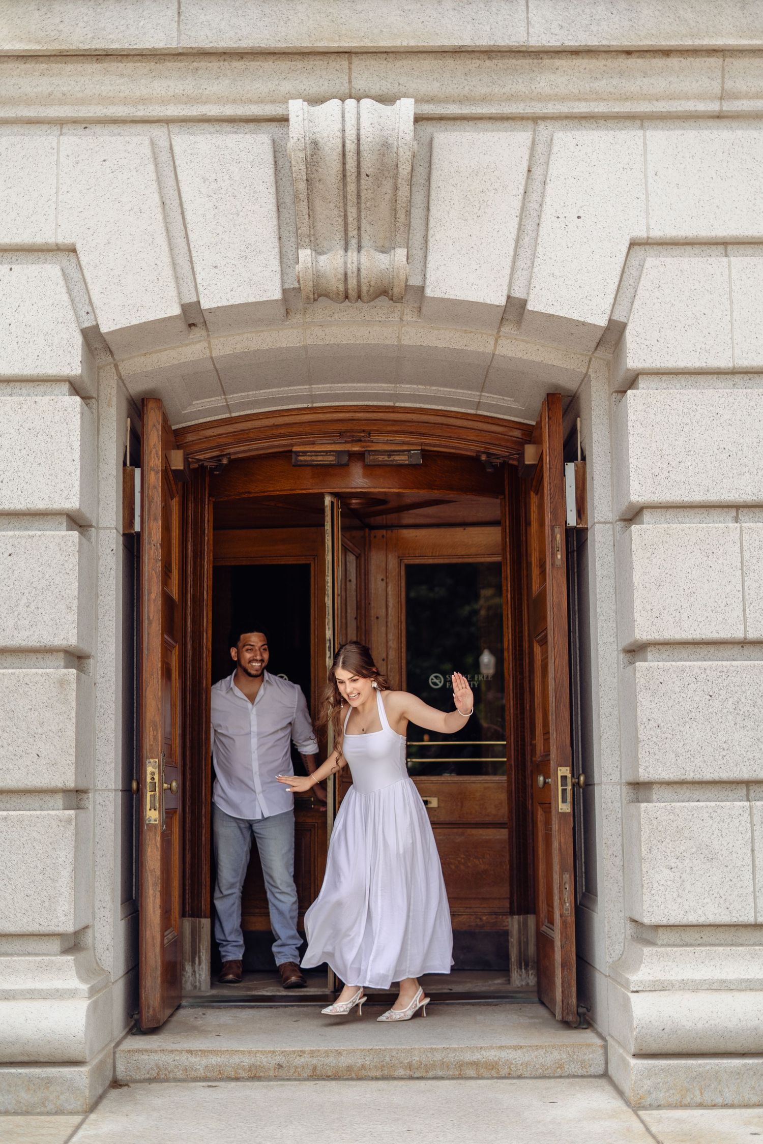 Couple exits elegant stone building with wooden doors, woman in white dress dancing, man in casual shirt smiling behind her.