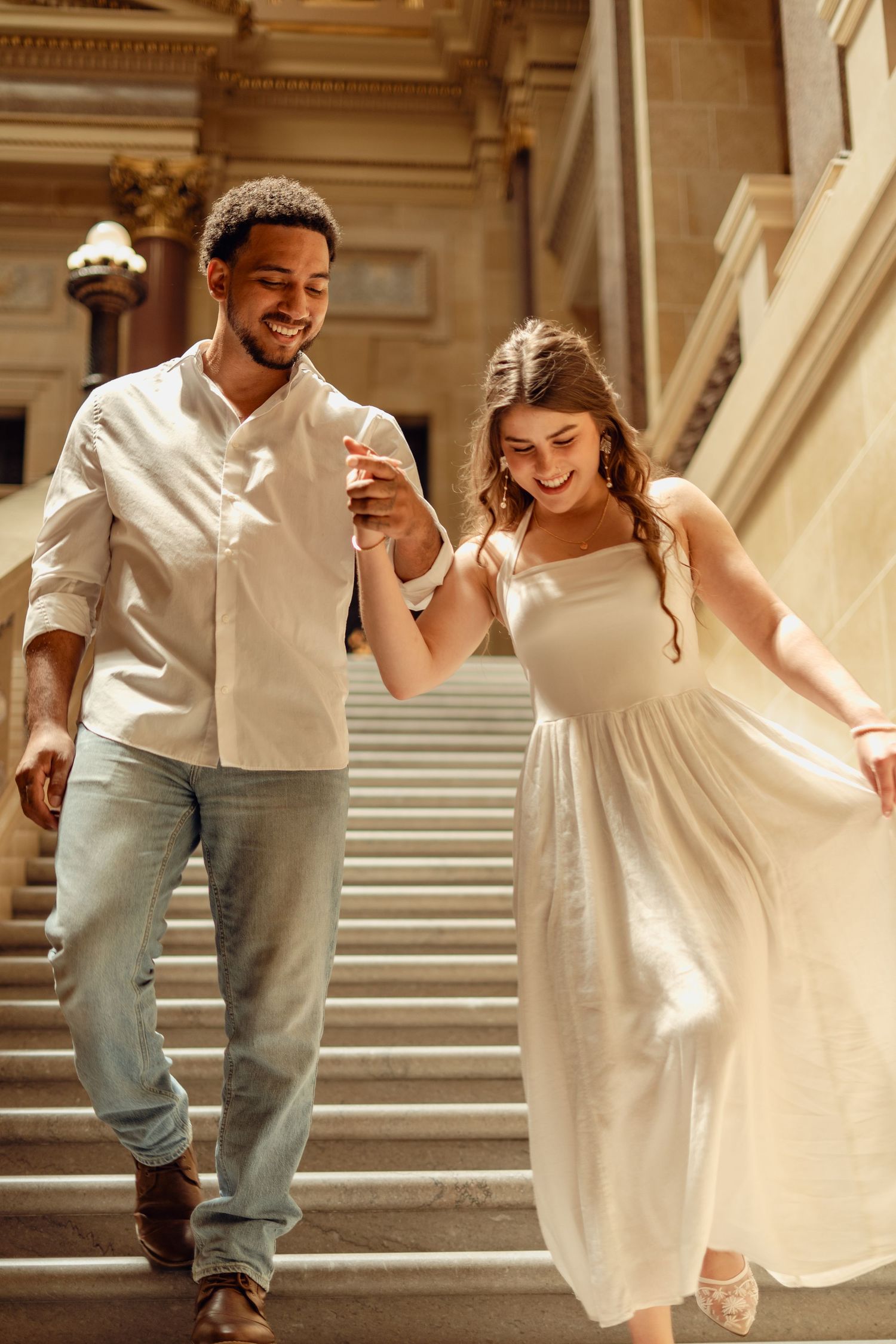 A happy couple holding hands and walking down grand stone steps, dressed in white outfits, smiling joyfully.