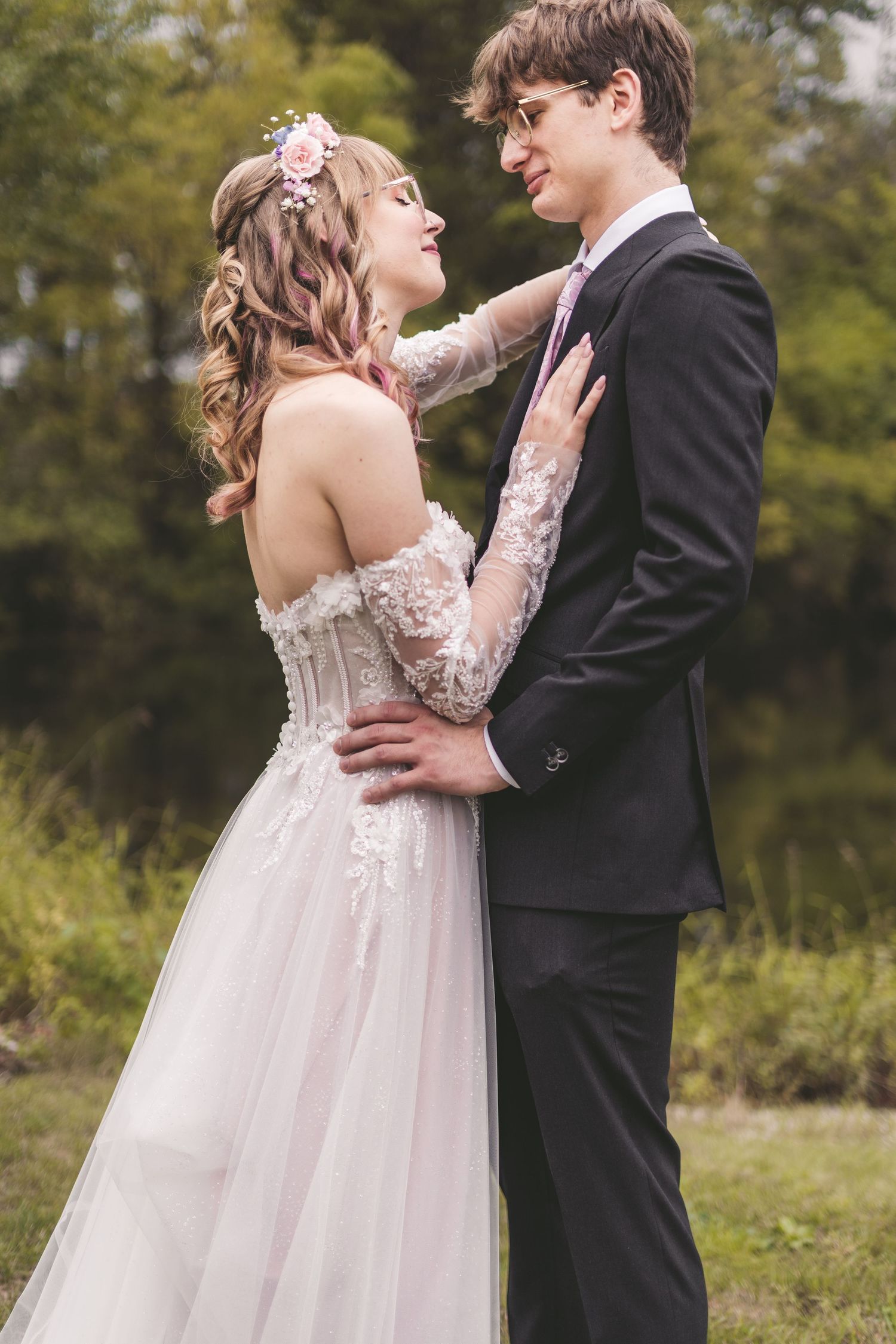 Bride and groom embracing outdoors, she wears off-shoulder lace gown with floral hair piece.
