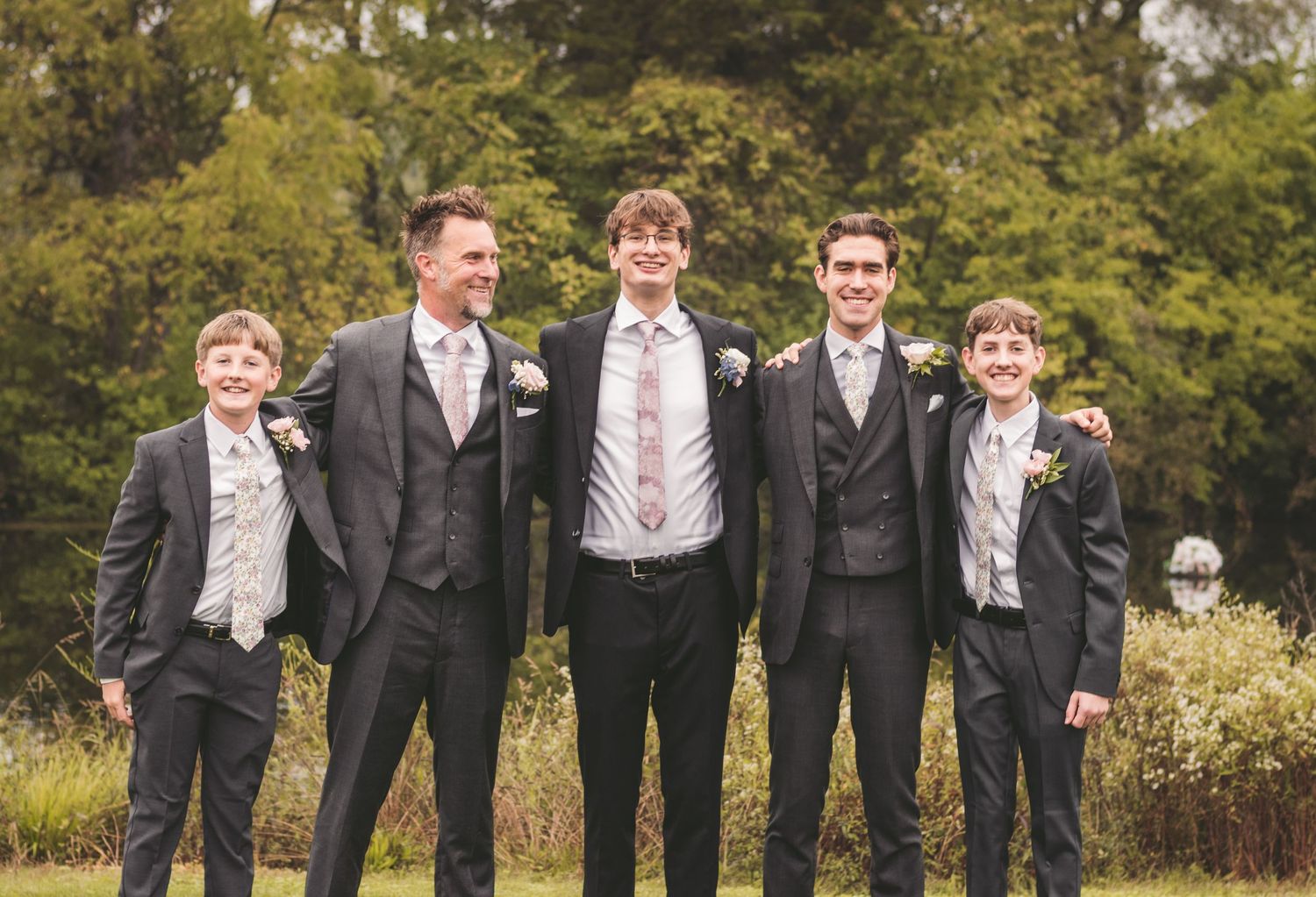Five males in matching grey suits posing together outdoors at a wedding celebration in a garden.