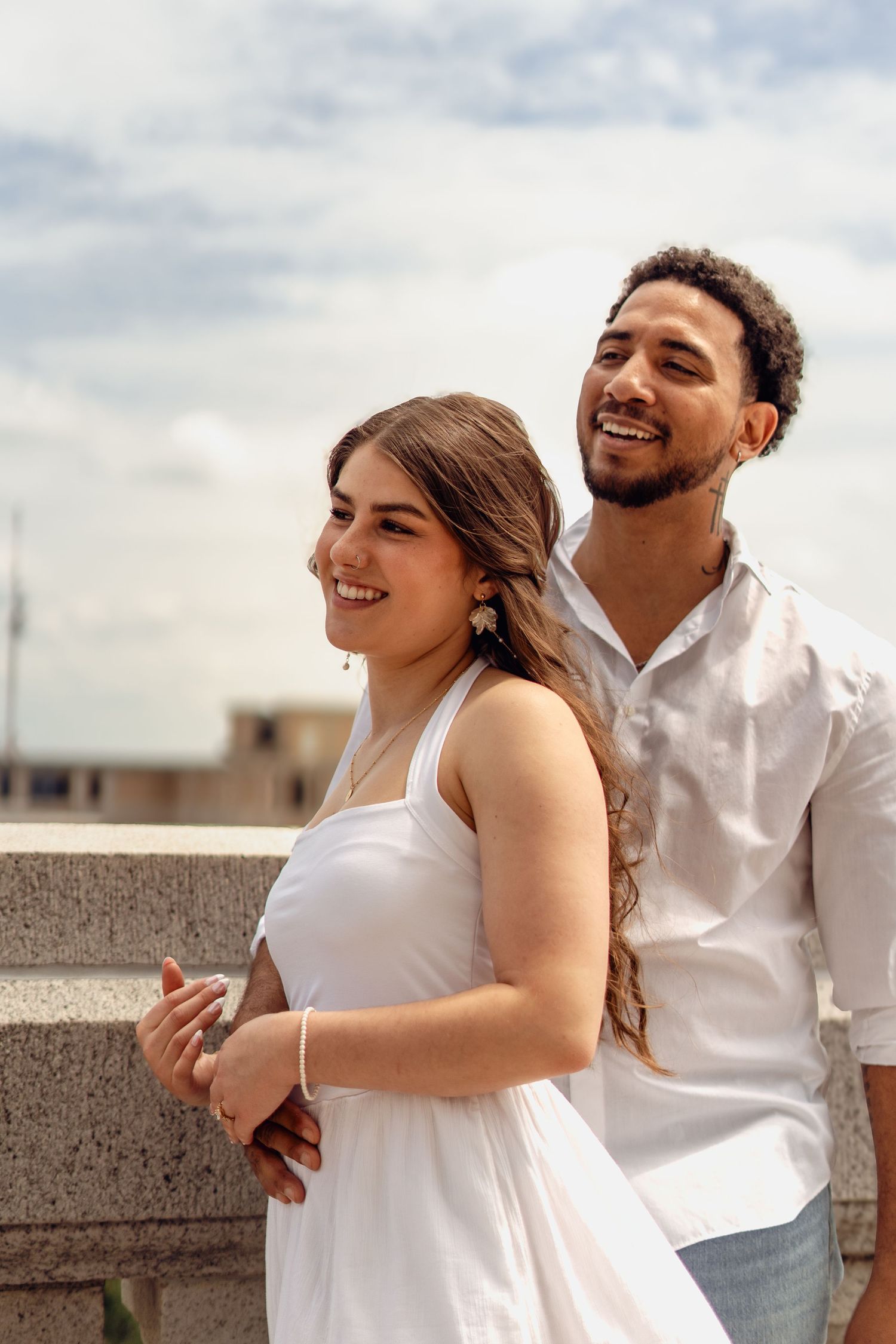 A smiling couple in white outfits poses outdoors against a cloudy sky, the man embracing the woman from behind.