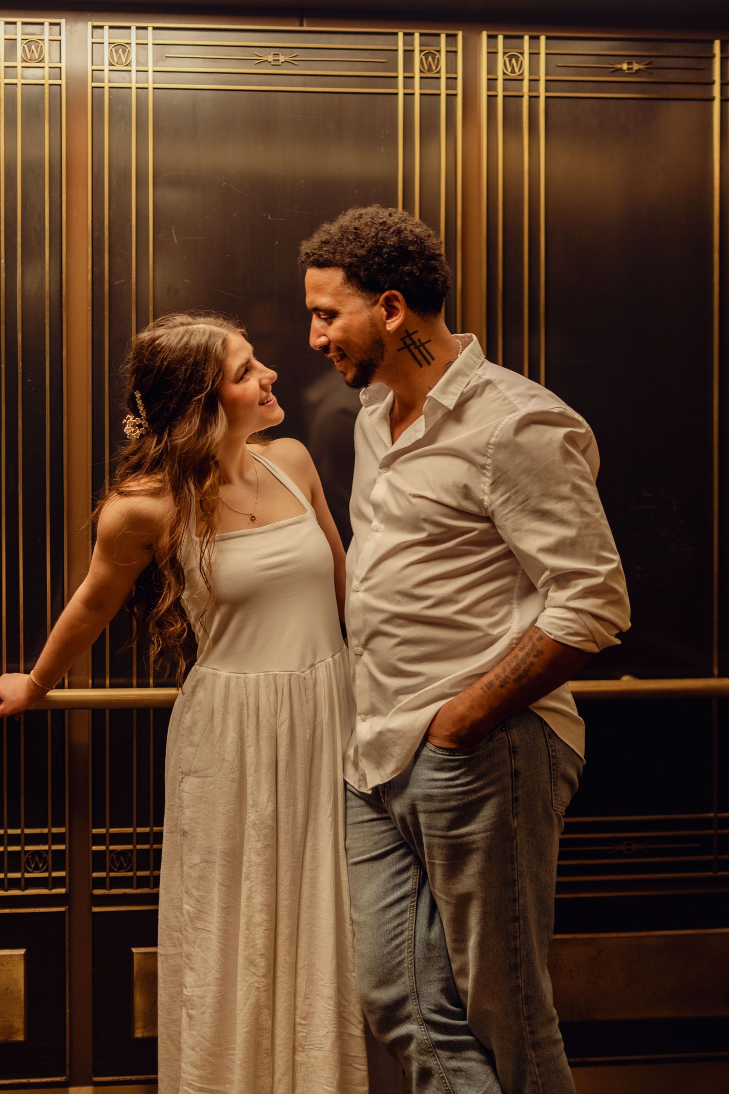 Romantic interracial couple gazing at each other in golden art deco elevator, woman in white dress and man in white shirt.