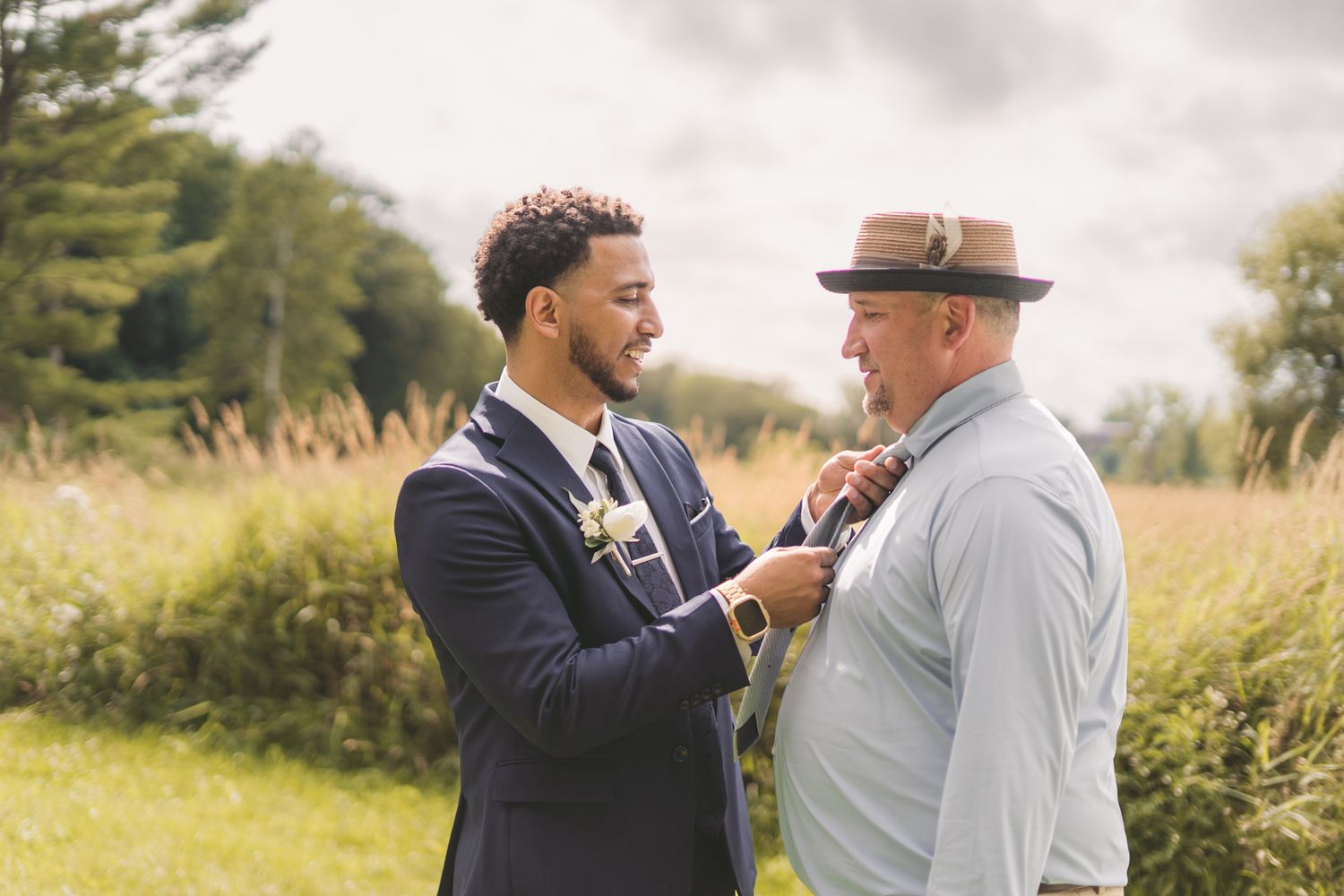 Two men in formal attire adjusting neckties outdoors at a wedding ceremony with green fields in the background.