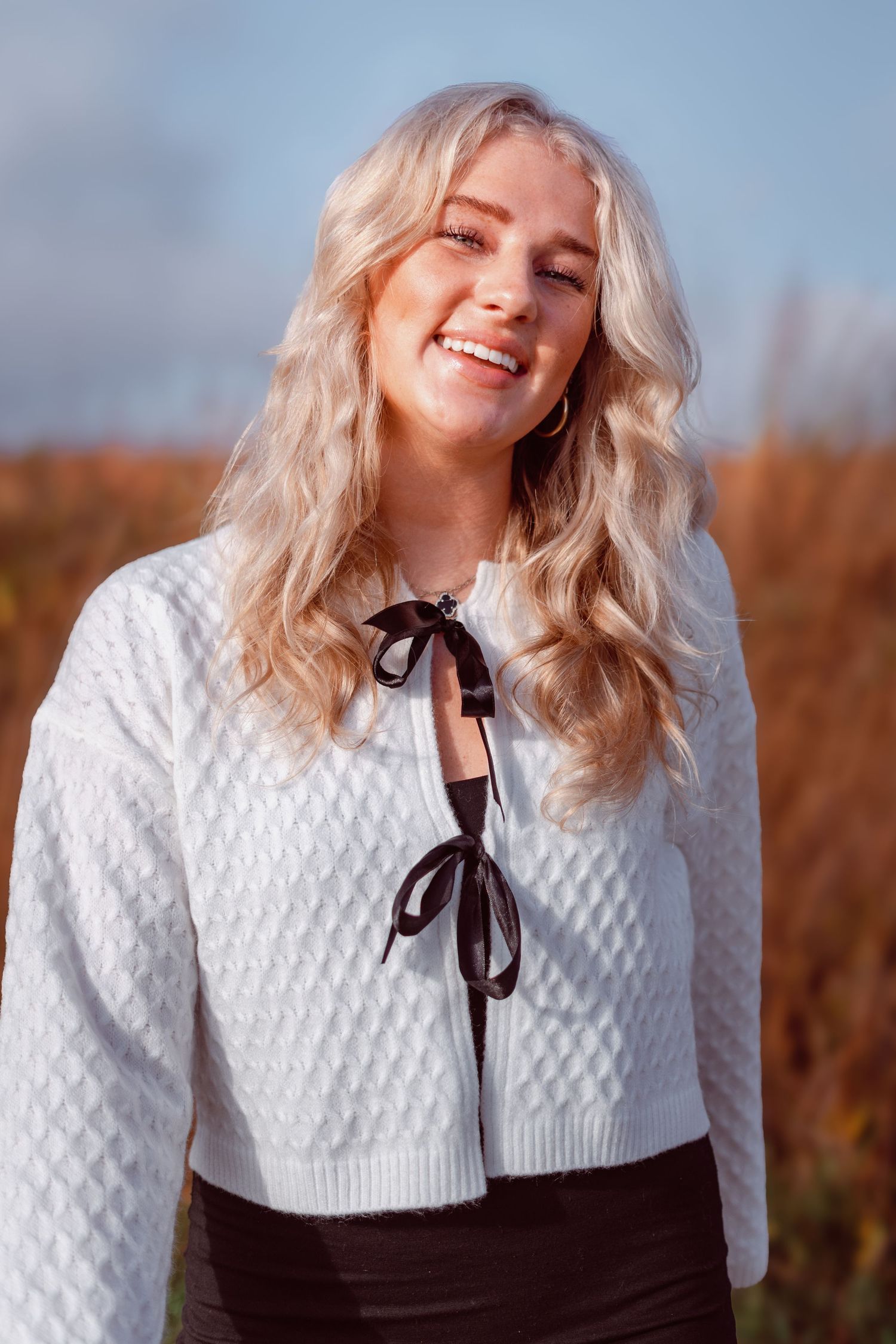 A person wearing a white knit sweater with a black bow tie detail smiles joyfully in an outdoor autumn setting.