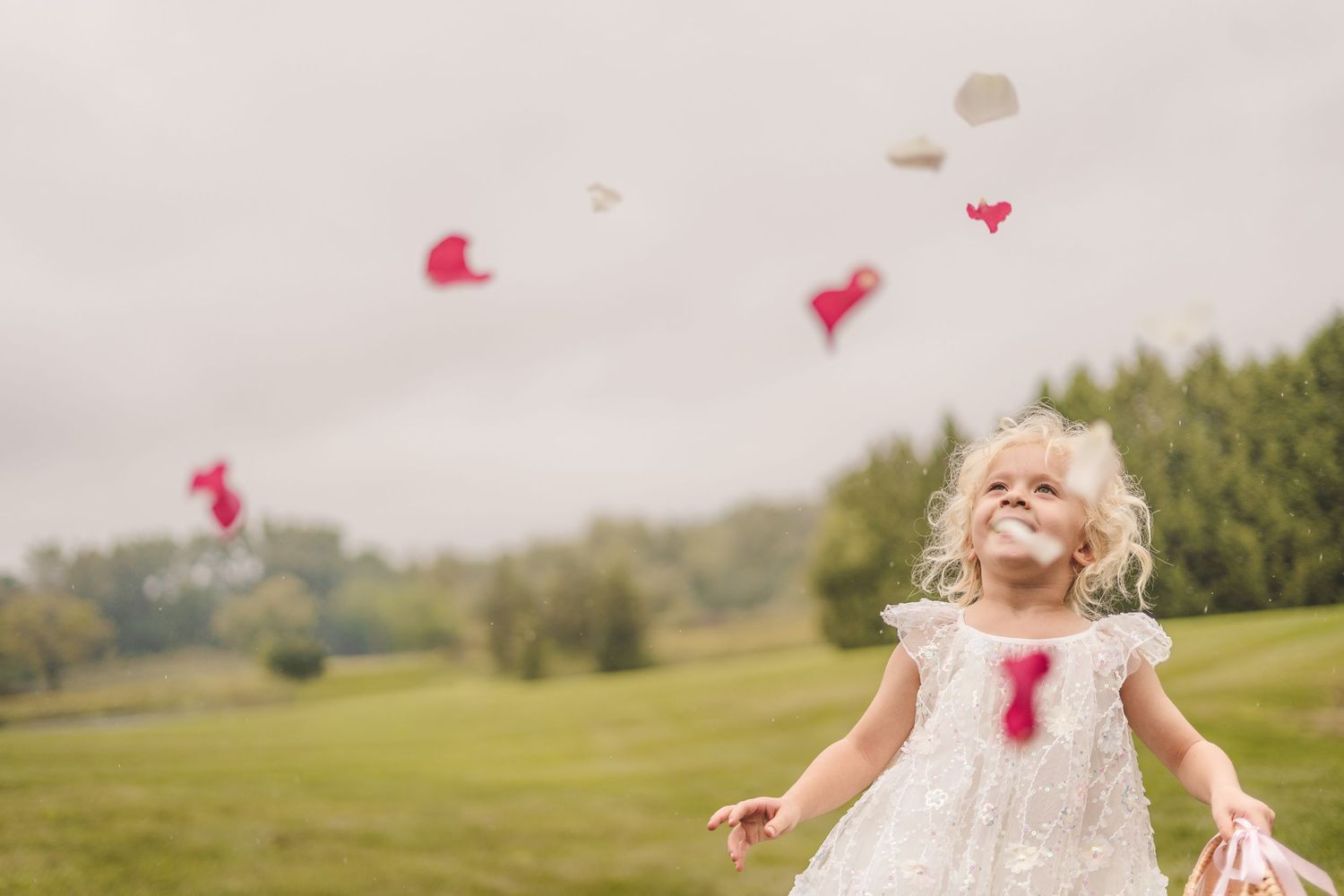 Joyful little girl in white dress tossing pink and white heart-shaped petals outdoors.
